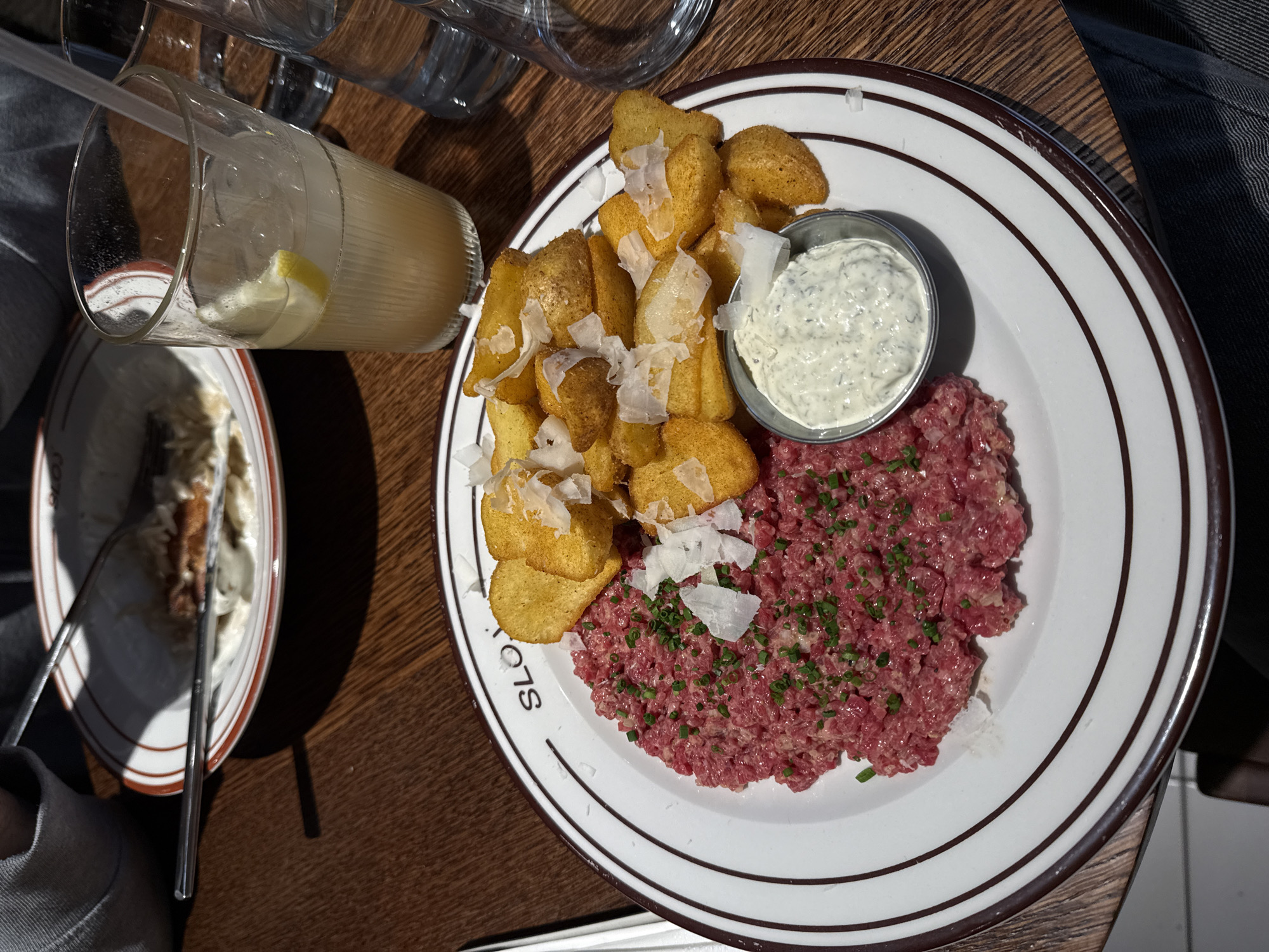 The image displays a close-up, overhead view of a meal served at a restaurant in Belgrade, Serbia, identifiable by the partial "SLO" branding on the main plate, consistent with "Slovenija" restaurant. The scene focuses on a round white plate with a brown double-line rim, placed on a dark brown wooden table.
The main dish on the plate consists of three distinct components: a mound of raw, finely ground red meat, likely beef tartare, garnished with chopped green chives on the left side; a pile of golden-brown potato wedges or robust chips on the right, topped with thin shavings of white cheese; and a small, silver metal ramekin centrally placed at the bottom, containing a creamy white sauce with visible green specks, resembling tartar sauce.
In the upper right portion of the frame, a tall, clear glass with a ribbed texture holds a pale yellow beverage with ice cubes, a lemon wedge, and a clear straw. Another clear glass, likely containing water, is partially visible to the right of the first glass.
In the upper left background, a partially consumed meal on a white plate with a brown rim is visible, appearing to contain a white creamy base with a piece of brown food and a metal fork or spoon resting within. A person's grey sleeve is partially visible next to this plate. The lower right edge of the frame shows a partial view of what appears to be a person's blue-grey denim-clad leg or lap. The overall lighting suggests an indoor setting, possibly with natural light from a nearby window or strong artificial illumination.