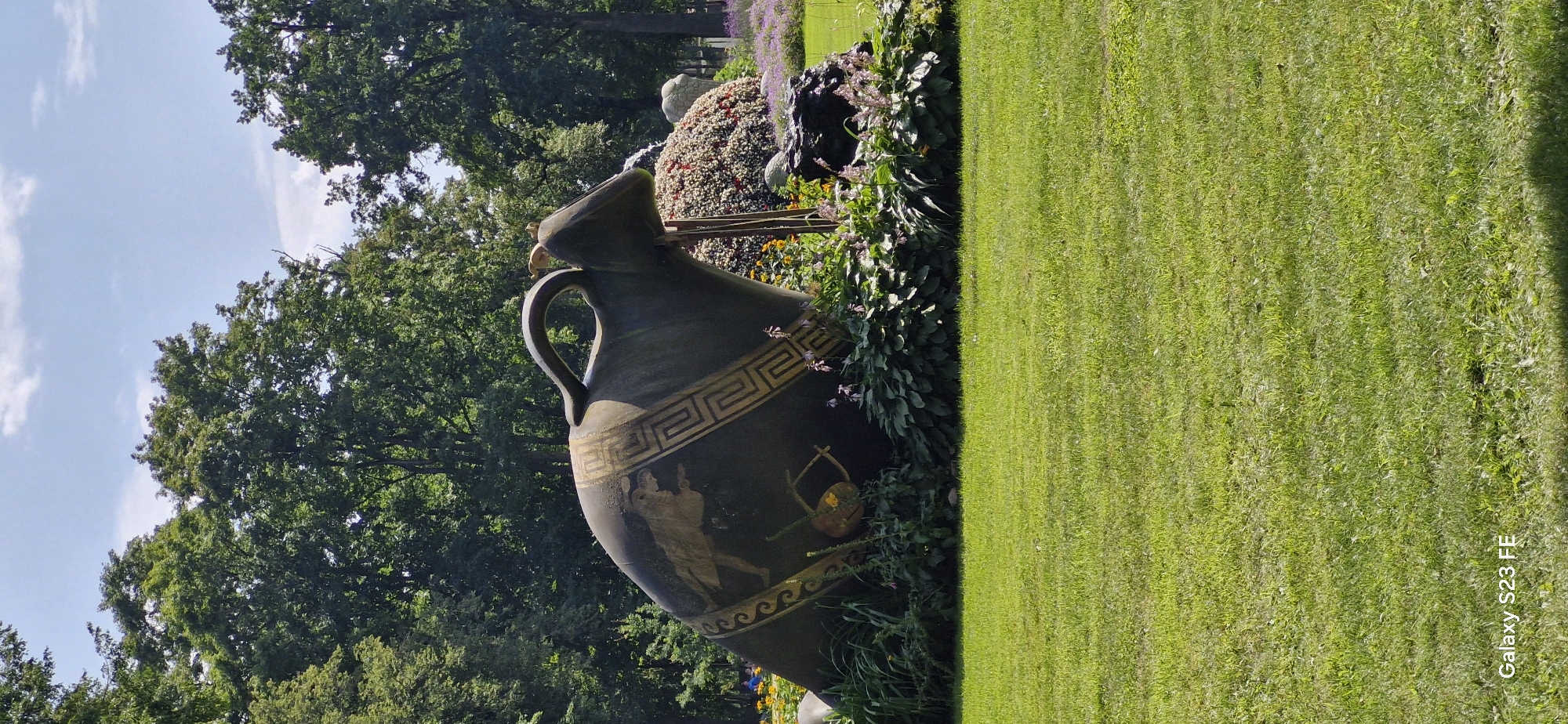 This is a vertical, eye-level, medium shot of an ornate amphora sculpture in a park setting. The amphora, dark green with gold detailing, is large and rests on its side, with its opening facing towards the right. It is decorated with a Greek key pattern and a depiction of a winged figure. The sculpture is surrounded by lush green foliage and some small purple and yellow flowers. In the background, there are mature trees with dense green leaves, and a bright blue sky with scattered white clouds. The foreground is a well-maintained green lawn. The lower left corner of the image shows the watermark "Galaxy S23 FE." The sculpture is located in the city of Pakruojis, Lithuania, specifically within the Pakruojis Manor park complex.