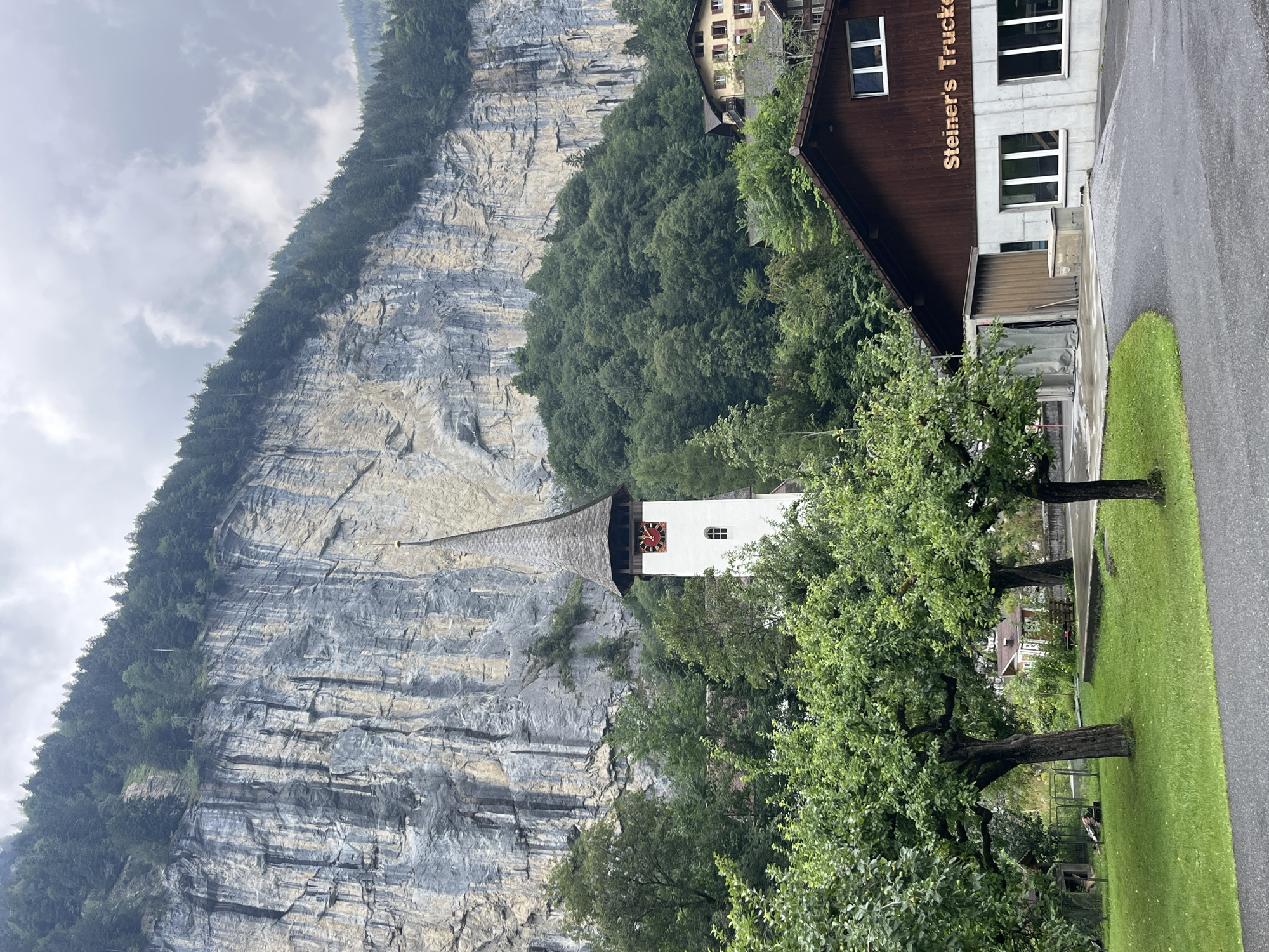 In Wengen, Switzerland, a dramatic scene unfolds against a backdrop of towering, sheer rock cliffs, partially obscured by lush green trees. In the foreground, a paved road curves around a well-maintained lawn with two mature apple trees. To the right, a two-story building with a dark wooden upper section and a concrete lower section stands, featuring large windows and the prominent signage "Steiner's Trucker." A tall, white church tower with a distinctive dark, pointed roof rises above the trees, its face adorned with a red clock. The overall environment suggests a village nestled within a formidable natural landscape, under a cloudy sky.