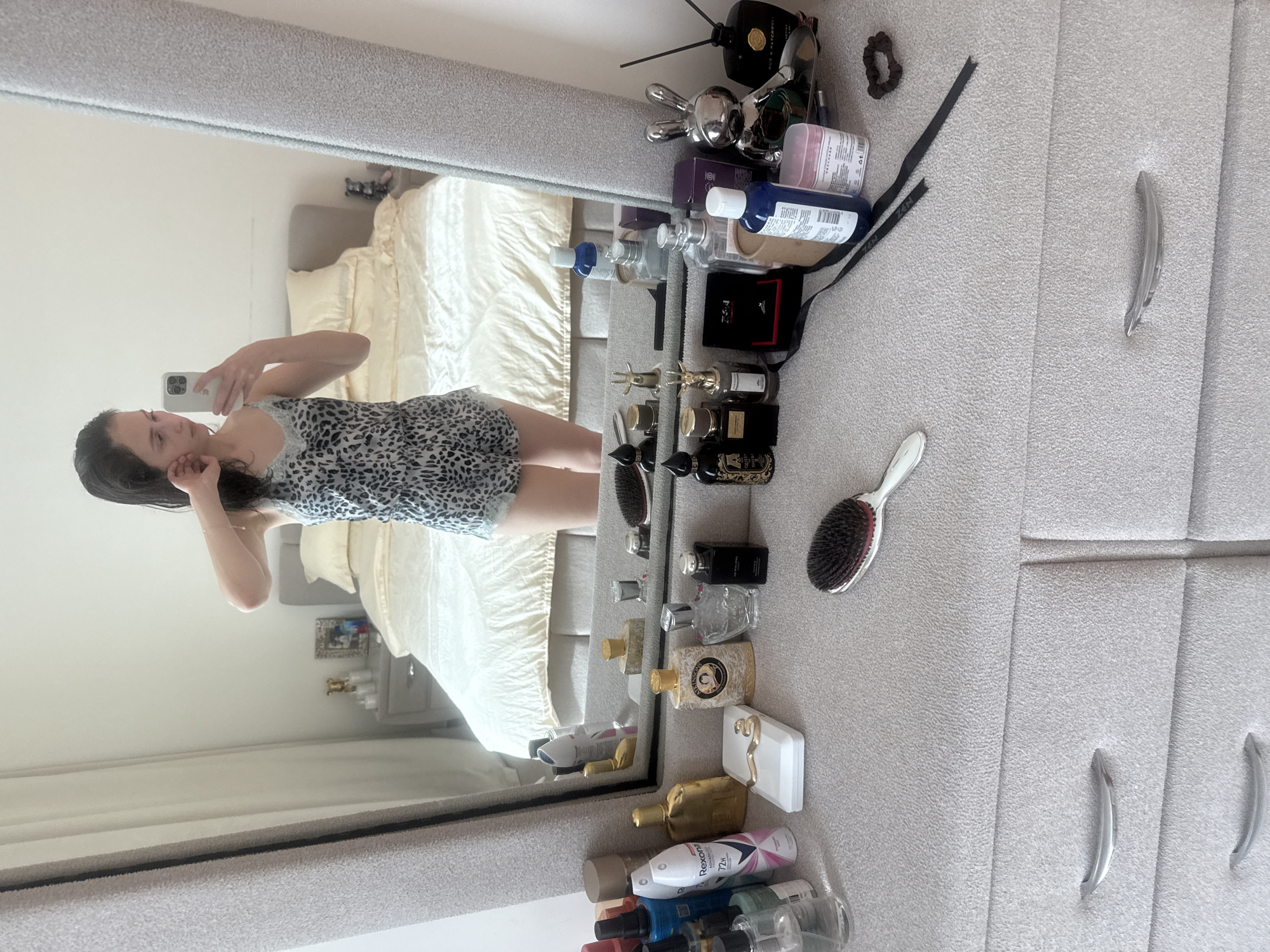 A woman is taking a mirror selfie in a bedroom. In the mirror, a double bed with light-colored bedding is visible. The woman is wearing a patterned tank top. To the right of the mirror, on a light-gray surface, are various toiletries and accessories, including perfume bottles, a hairbrush, and hair ties. The background includes a wardrobe with metal handles. It is not possible to determine if the location is in the city of Abu Dhabi, United Arab Emirates, from this image.