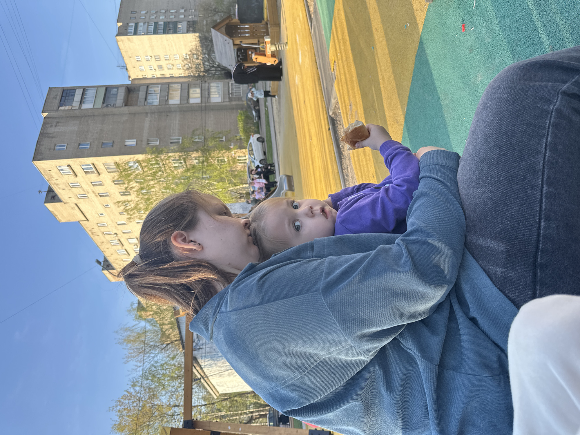 A young woman with her hair in a ponytail is holding a small child. The woman wears a blue jacket and the child wears a purple shirt and holds a piece of bread. The woman appears to be kissing the child's head. They are outdoors in a playground with a yellow and green surface. In the background are apartment buildings, parked cars, and other people. The location may be Tomilino, Russia, based on the context.