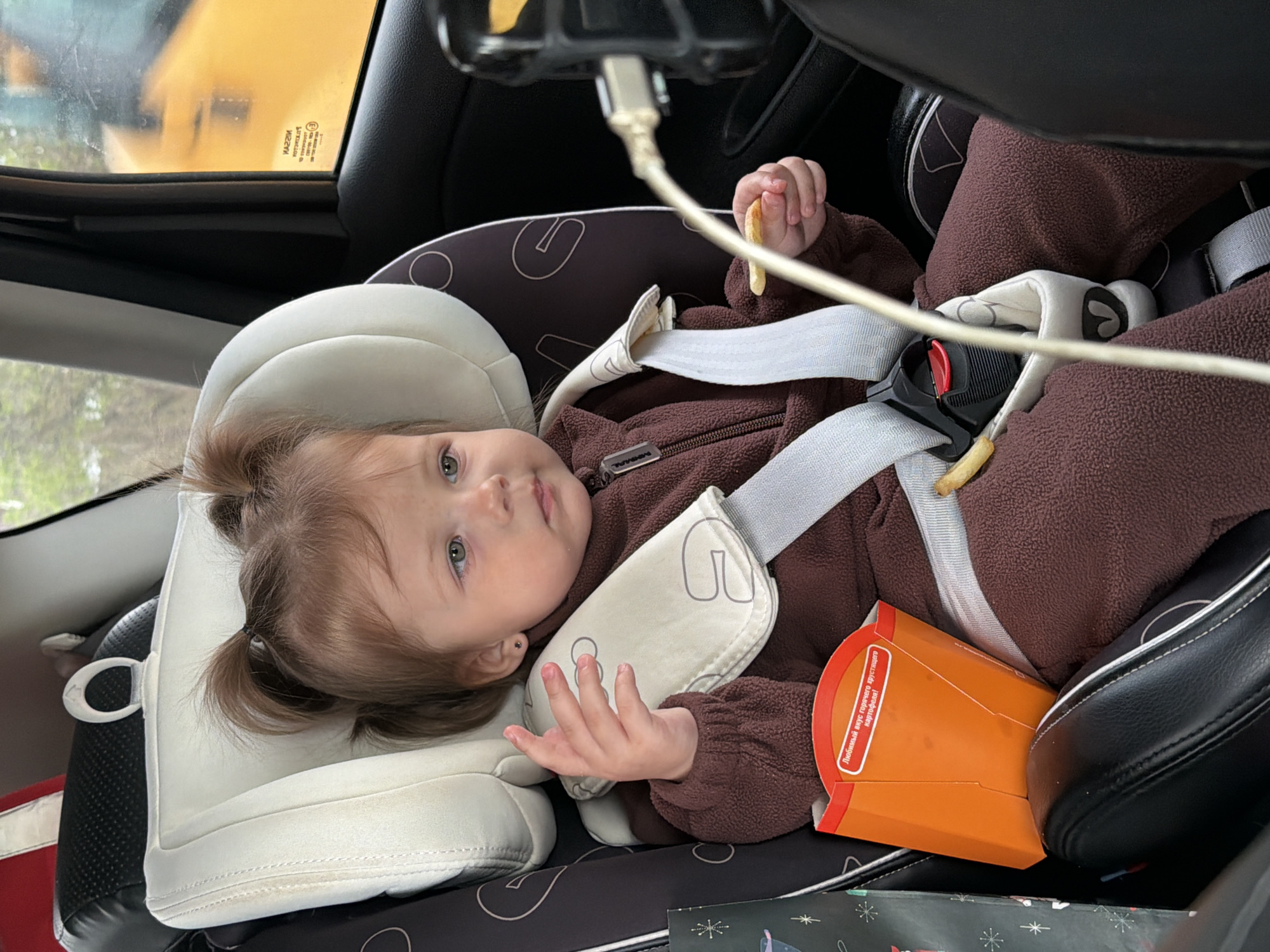 A young child is secured in a car seat inside a vehicle. The child wears a brown fleece suit and is strapped in with white harness restraints. The child has pigtails and is holding a french fry in one hand. An orange cardboard container, presumably holding more french fries, sits beside the child on the car seat. A phone charging cable is visible near the top of the frame. The location is implied to be Ширяево, Russia based on the fact you told me to specify that location.