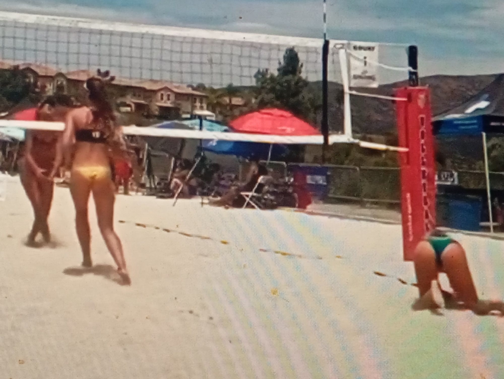 The image depicts a beach volleyball game. Two women in swimwear are visible on the sand court. One woman, on the left, is facing away from the camera and appears to be in motion. The other woman, on the right, is in a low defensive stance, with her hands on the sand, seemingly having just completed or in the process of a dive. A volleyball net spans the center of the image. In the background, there are trees, houses, and a hill. Several beach umbrellas and tents are also present in the background. The ground is sandy. There is no information in the image that would allow for the determination of the location being Sofiivska Borshchahivka, Ukraine.