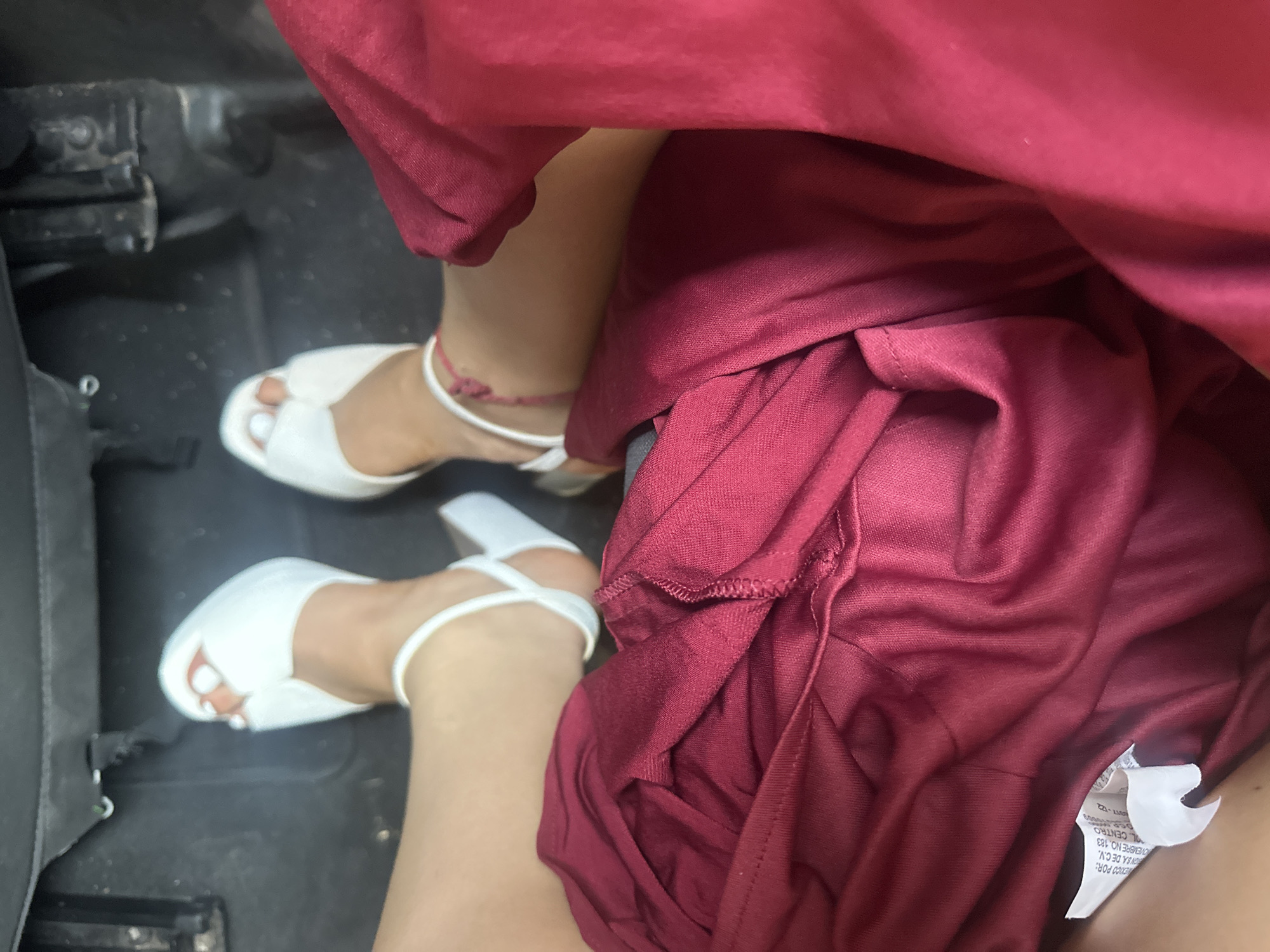 The image shows a person sitting inside a vehicle, presumably a car, wearing white high-heeled sandals and a burgundy dress. The setting appears to be the interior of a vehicle, with the person's feet and lower legs visible. The dress tag has some text in Spanish and mentions "OVEMBRE NO. 183", and the phrase "L. CENTRO" suggesting that the dress might be purchased from a shop in "Centro" (Central area) of the city of Puebla, Mexico.