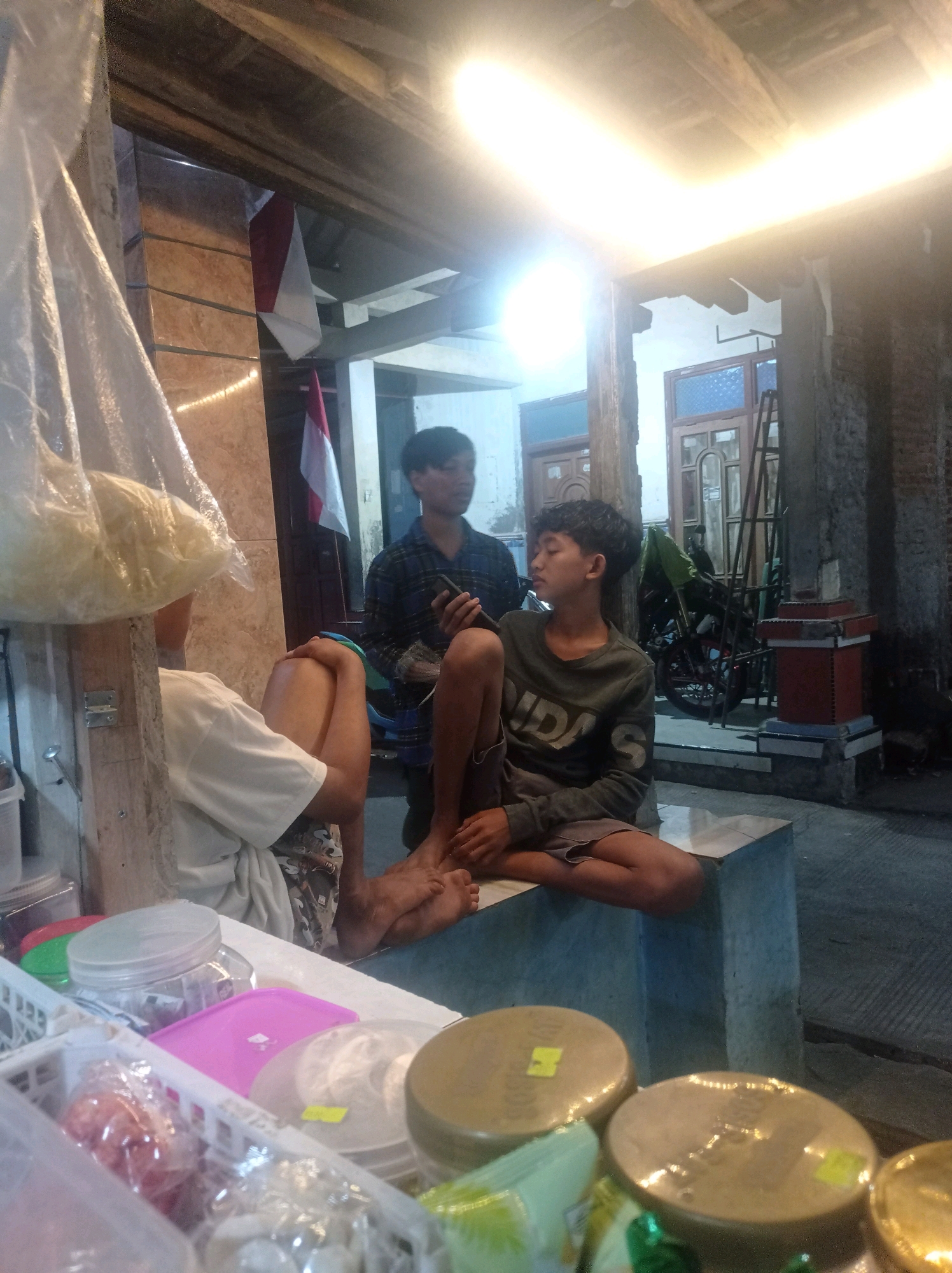 The image depicts an outdoor scene in the city of Babalan, Indonesia, likely a roadside stall or shop at night. Two young men are the primary subjects. One is seated on a raised concrete platform, looking to the side with his eyes closed, wearing a dark long-sleeved shirt with a white logo and grey shorts. He is barefoot. The other young man stands behind him, looking down at a mobile phone he holds in his right hand, wearing a checkered blue and black long-sleeved shirt. Partially visible in the foreground is another person wearing a white t-shirt and patterned shorts, with their legs crossed and feet visible. Various items are present, including plastic bags filled with food items, plastic containers, and what appear to be large circular food products wrapped in plastic on a counter. Indonesian flags are visible in the background. The environment appears to be dimly lit, with a bright artificial light source overhead casting strong highlights. The structures suggest a simple, possibly temporary, setup common in local markets or informal businesses.