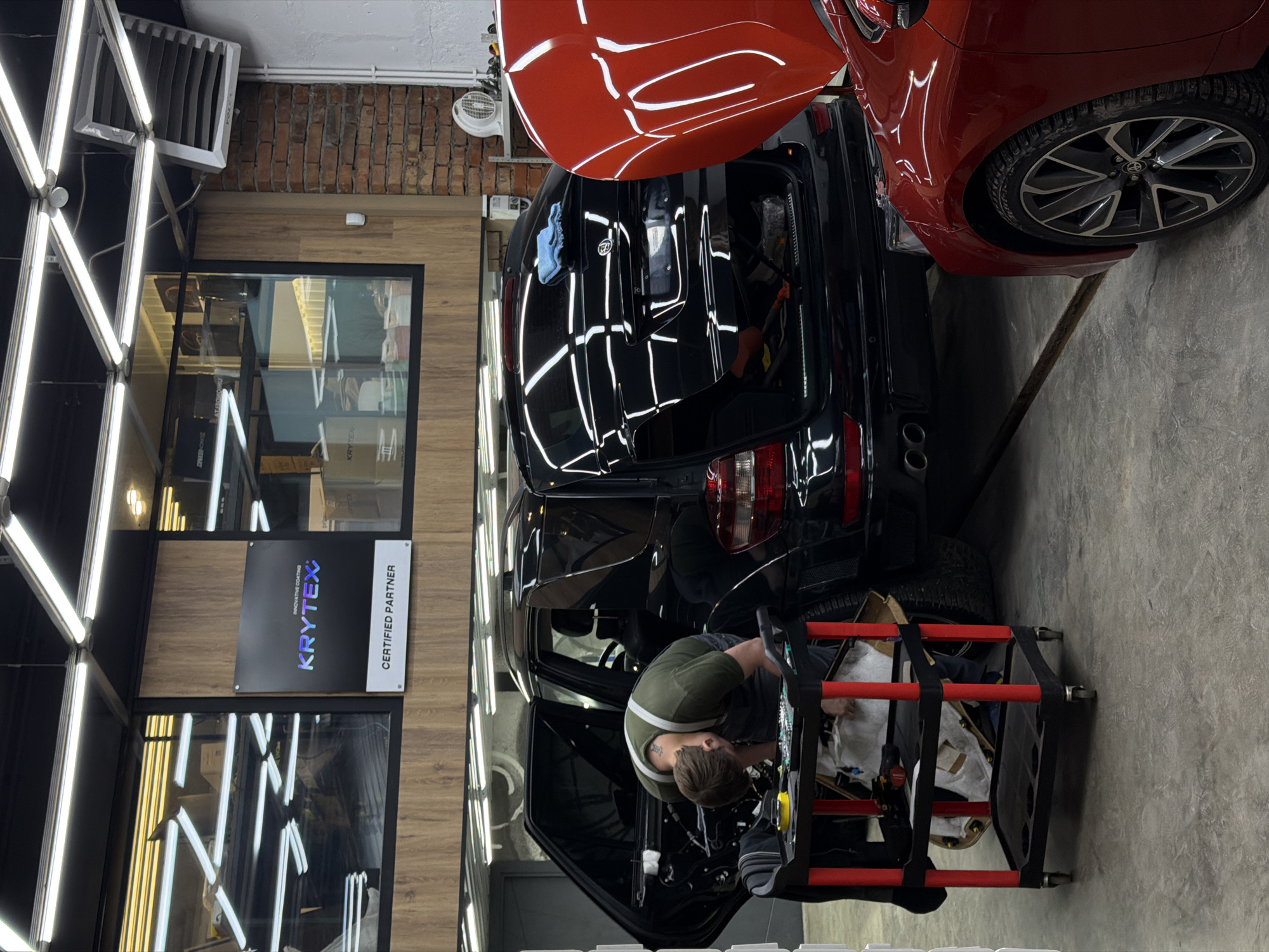 A man wearing a green shirt and an apron is working on the interior of a black Mercedes-Benz in what appears to be a car detailing or repair shop. The trunk of the Mercedes is open, and a red car is parked nearby. The shop has bright lighting fixtures on the walls and ceiling, a wooden paneled wall with a "Krytex Certified Partner" sign, and a concrete floor. The location is likely in the city of Krasnoyarsk, Russia, based on the presence of a Krytex Certified Partner, a Russian brand specializing in automotive care. A red and black rolling tool cart is positioned near the open door.