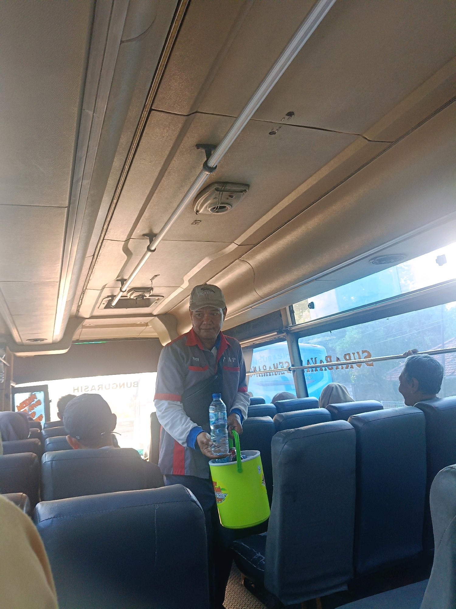 An elderly man wearing a grey shirt with red and white trim and a grey baseball cap with the word "JAGUAR" on it is standing inside a bus. He is holding a clear plastic bottle of water and a bright green bucket with a handle in his right hand. The man is looking at the camera with a slight smile. The interior of the bus has rows of dark blue seats. Through the windows, trees and a building can be seen, suggesting an outdoor environment. The bus appears to be in motion, indicated by the lighting and the slight blur of the background. The text "BUNGASIH" is visible on a sign in the background, which is likely the name of the bus company or a destination. The scene is set on the interior of a bus in Kedungrejo, Indonesia, likely a public transportation vehicle.