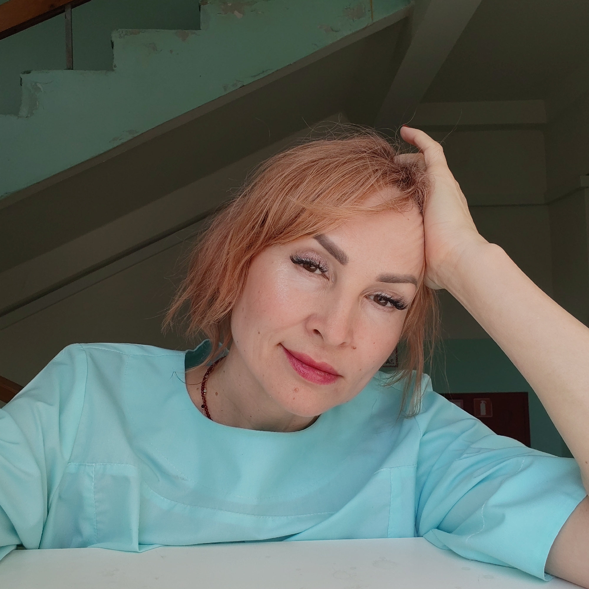 A woman with short, reddish-blonde hair is resting her head on her hand, looking towards the camera. She has fair skin and is wearing a light blue collared shirt. A beaded necklace is visible around her neck. The background includes a faded light green wall with what appears to be a staircase. Based on the environment and information from the user, this image was taken in Chaikovsky, Russia. A door and signage can be seen on the right side of the background. The woman is positioned at what appears to be a table.