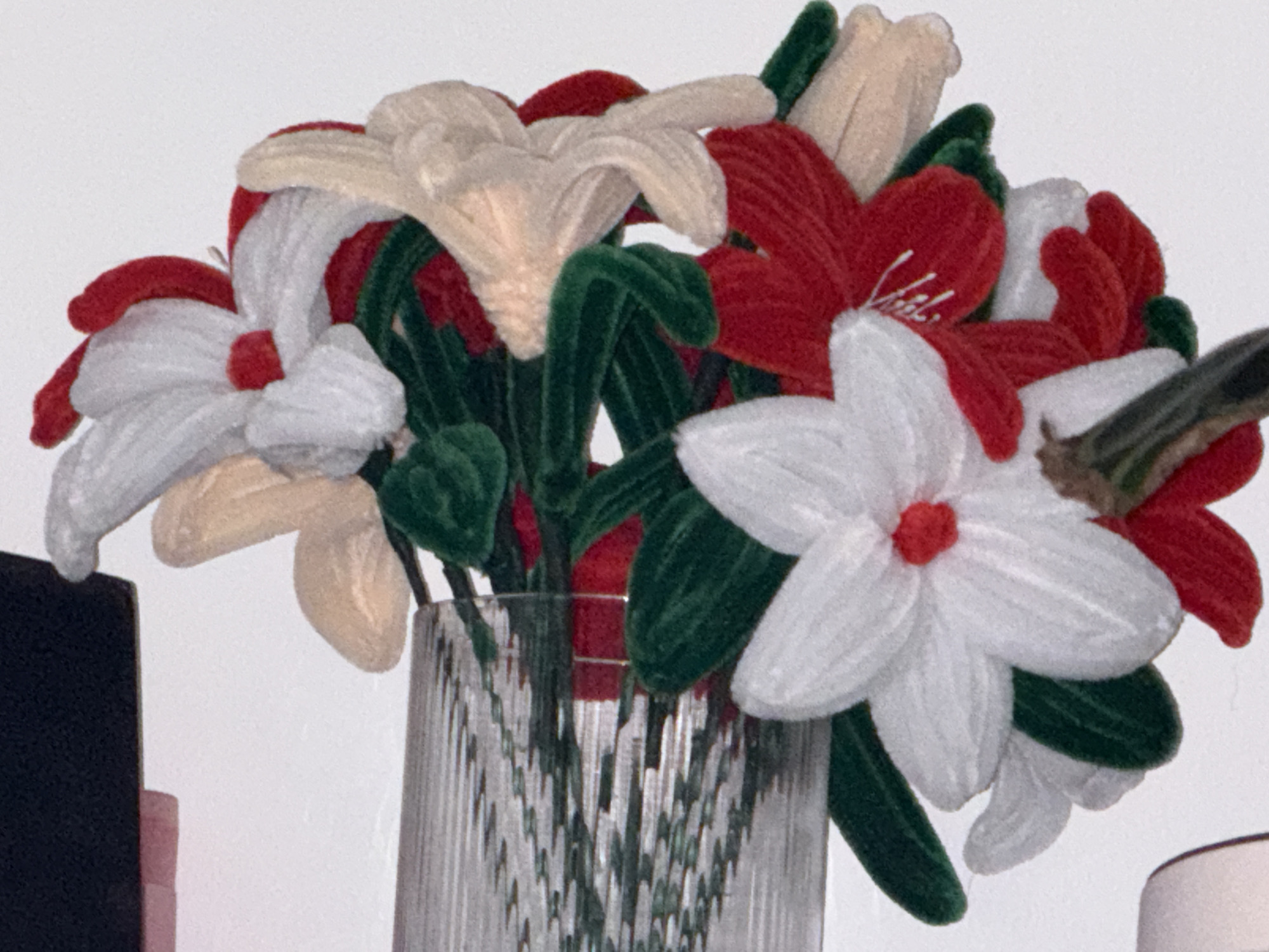 The image shows a close-up of a bouquet of artificial flowers made of pipe cleaners and felt. The flowers are a mix of red, white, and cream colors, with green stems. They are arranged in a clear glass vase with a ribbed texture. The bouquet is positioned against a white background. The context provided about Toholampi, Finland is not verifiable from the image.
