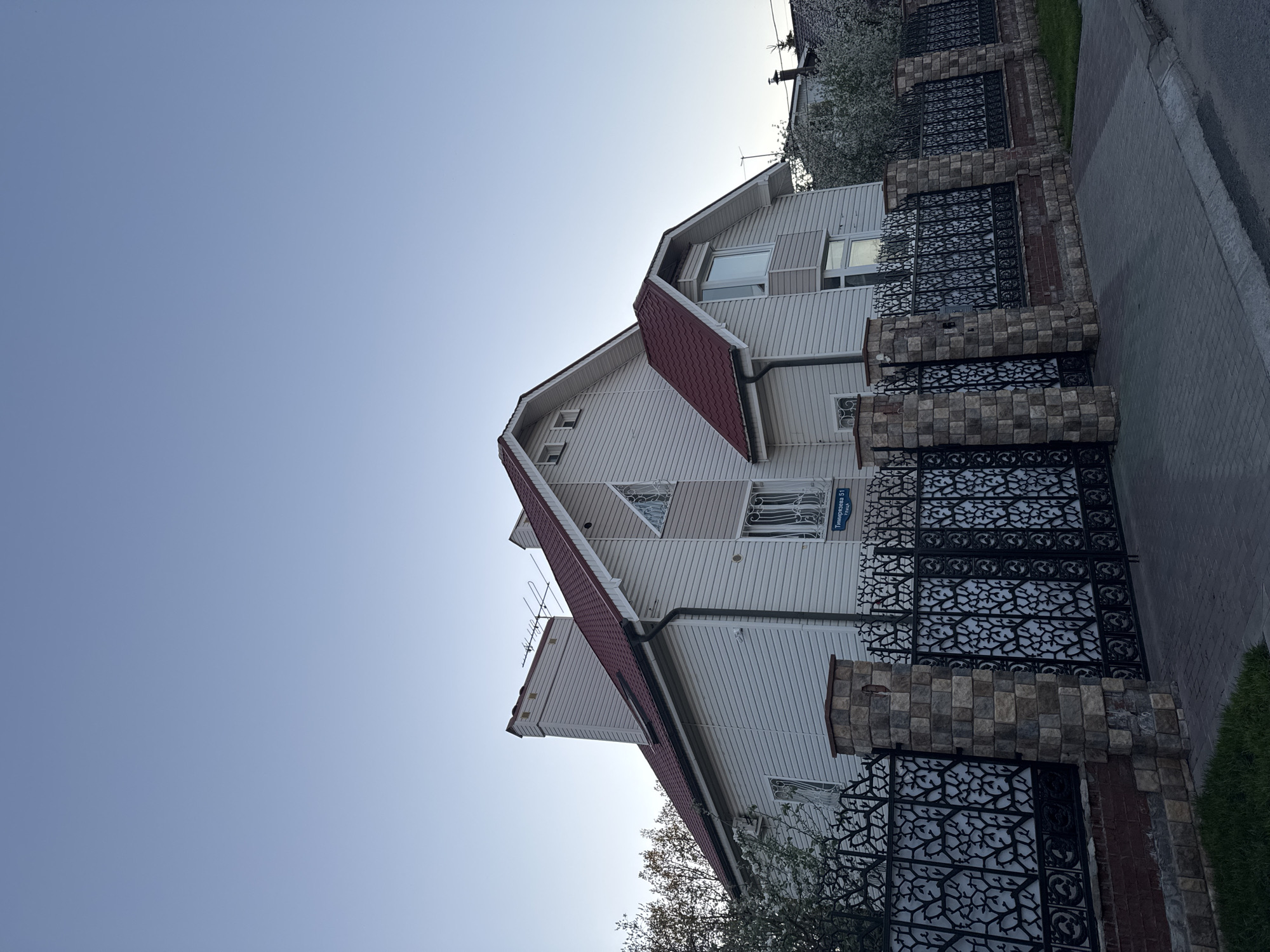 A low-angle shot shows a two-story house with a red roof and light siding in Unknown, Unknown. The front of the house faces the viewer, and a portion of the front yard, bordered by a decorative black metal fence and stone pillars, is visible. The sky is a clear, pale blue. A street sign reading "Тимирязева 51 улица" (Timiryazeva 51 street) is attached to the house near the fence. Trees with white blossoms partially obscure the house and fence in the foreground. A section of sidewalk is visible on the right side of the image.