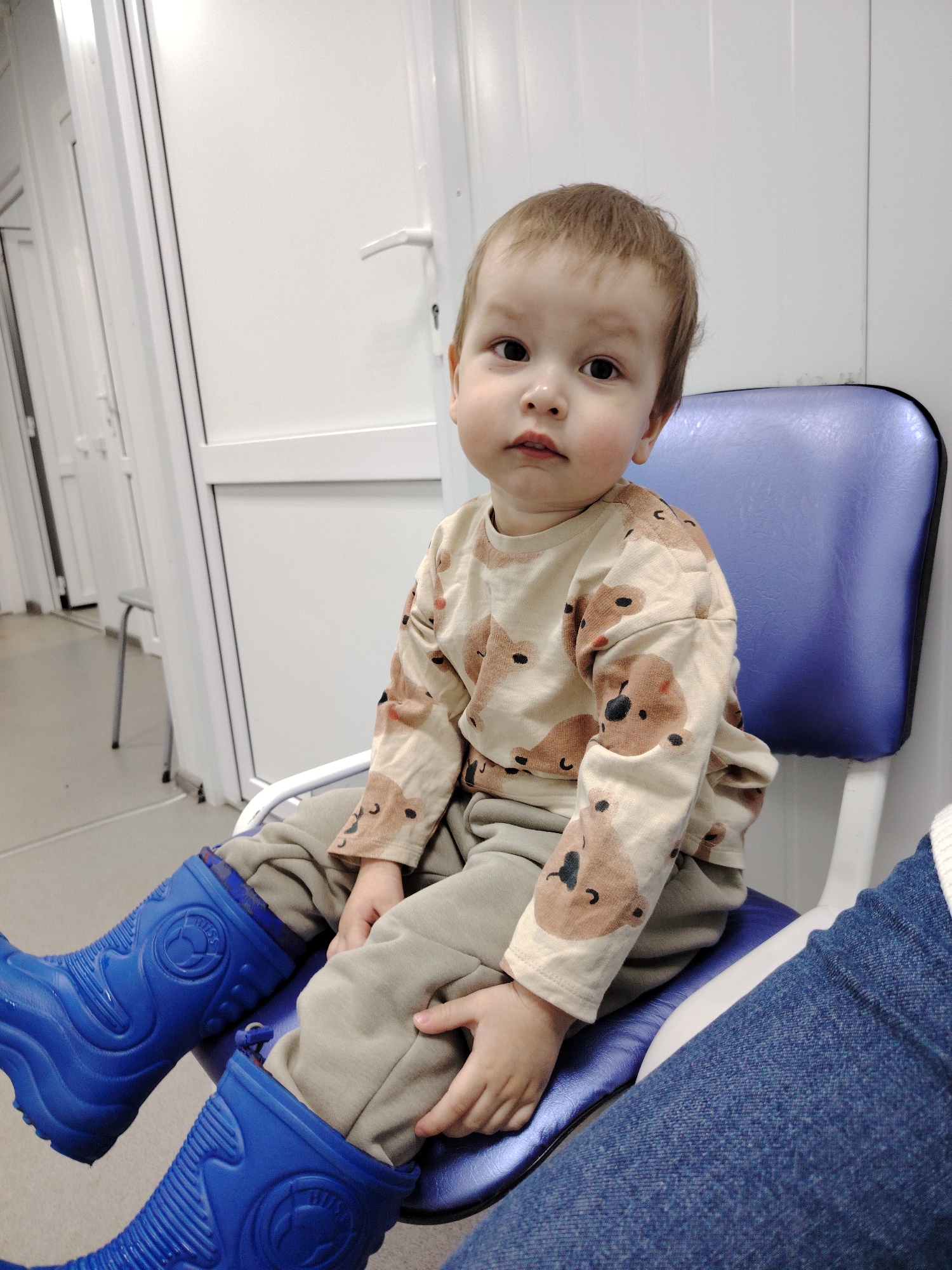 A young child sits on a blue chair, wearing a beige shirt with teddy bear prints, gray pants, and bright blue rain boots. The child is looking directly at the camera with a slightly apprehensive expression. The setting appears to be a sterile, white room, possibly in a clinic or hospital, indicated by the white walls and doors with silver handles in the background. A leg wearing denim jeans is visible to the right of the child. The location is in the city of Unknown, Unknown.