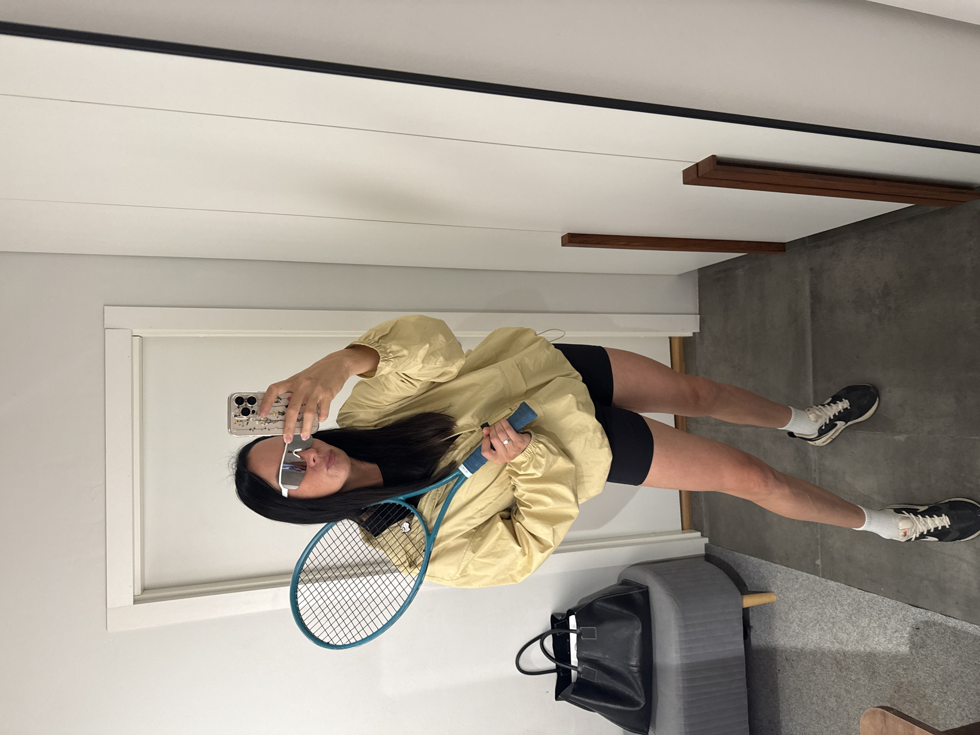 A woman is standing in a room, possibly a fitting room, holding a tennis racket and taking a selfie in a mirror. She is wearing sunglasses, a light yellow jacket, black shorts, white socks, and black and white sneakers. To her left, a black tote bag sits on a grey ottoman. The room appears to be sparsely decorated, with a white door and wall visible in the background. The floor is tiled and a gray rug is partially visible. The image does not provide any information about the city of Chaikovsky, Russia.