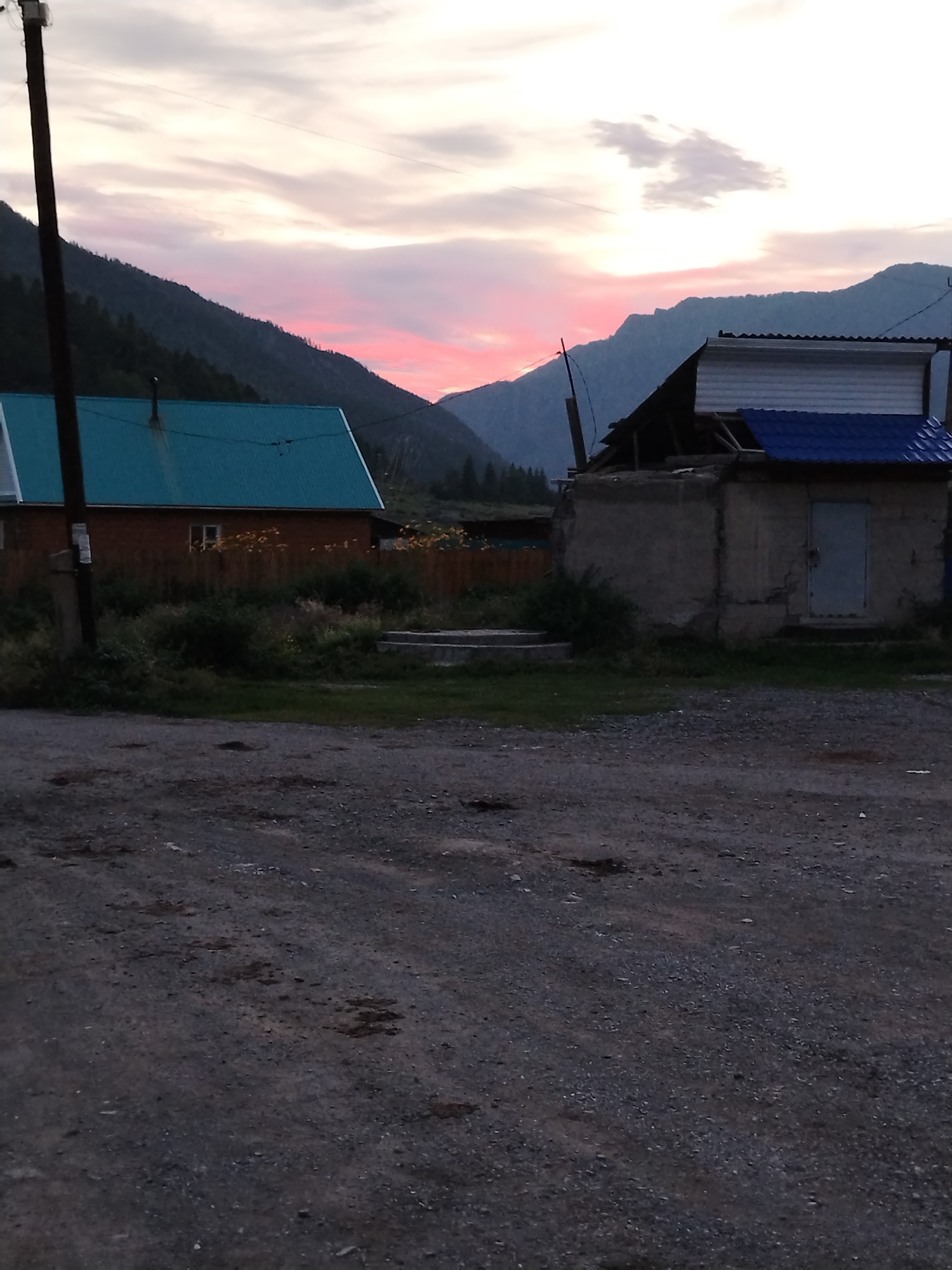 The image depicts a rural scene at dusk, likely in Iodro, Russia, with a mountain range in the background under a sky colored with hues of pink and grey. In the foreground, a gravelly yard is visible. To the left, a house with a teal-colored roof and wooden walls stands behind a wooden fence. To the right, a small, unfinished concrete structure with a blue-tiled roof and a gray door is present. A utility pole with wires is situated to the left of the teal-roofed house. Green vegetation and some steps are present between the yard and the houses.