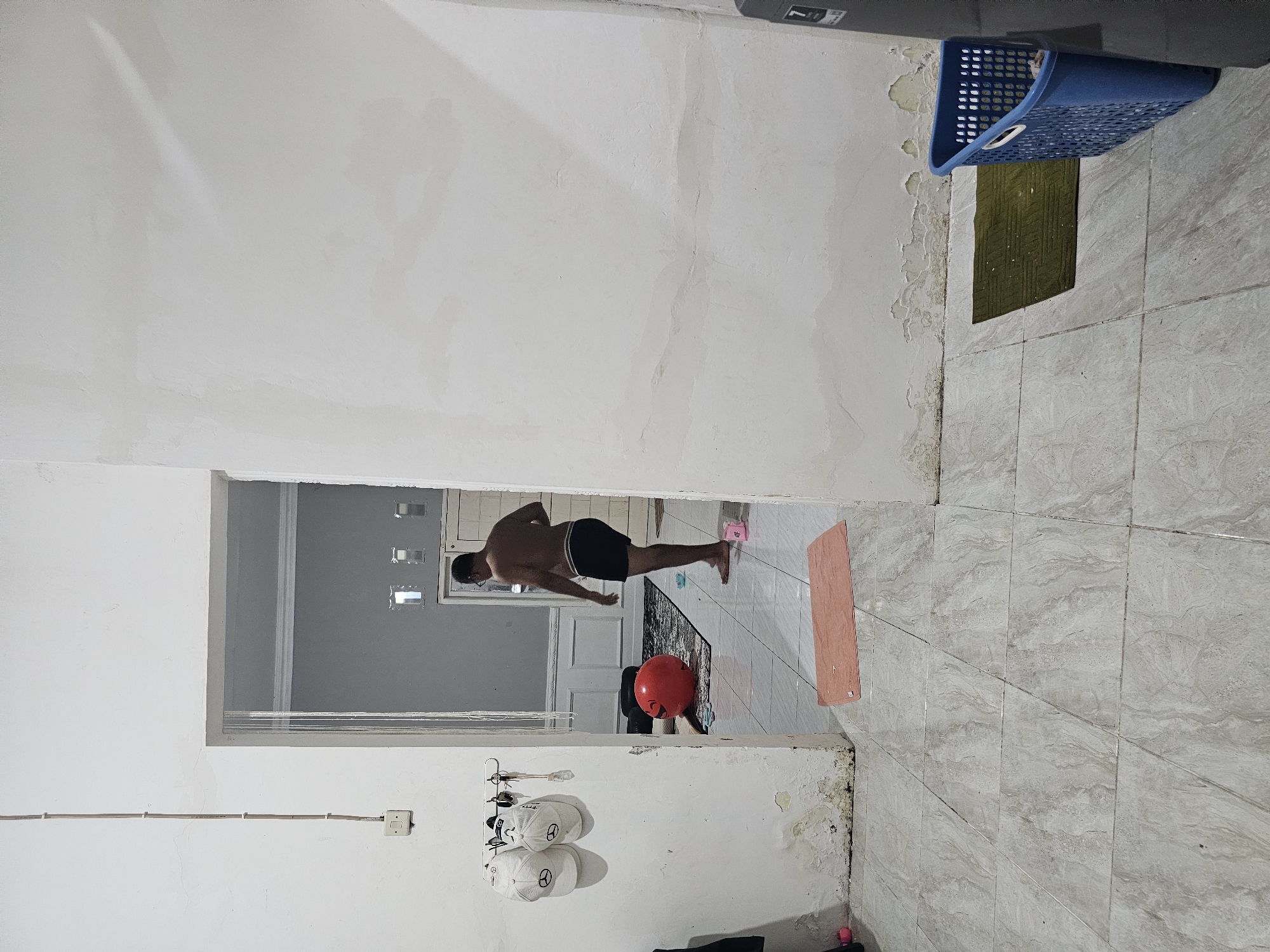 The image depicts an interior scene, possibly a residential hallway or room in Lamongan, Indonesia. A man, partially undressed and wearing black shorts, stands in a doorway, facing away from the camera and looking into an adjacent room. The room he is facing has a dark rug on the floor and a large, red, inflatable ball.
In the foreground, to the left of the doorway, two white baseball caps hang on a hook mounted on a white wall. Keys are also attached to the hook. The wall shows signs of water damage and peeling paint near the floor. The floor itself is tiled with a grey, marbled pattern.
To the right of the doorway, the white wall continues, showing similar signs of water damage. A green mat is placed on the floor, partially covered by a blue laundry basket. The overall environment appears to be a casual, possibly unfinished or undergoing renovation, living space.