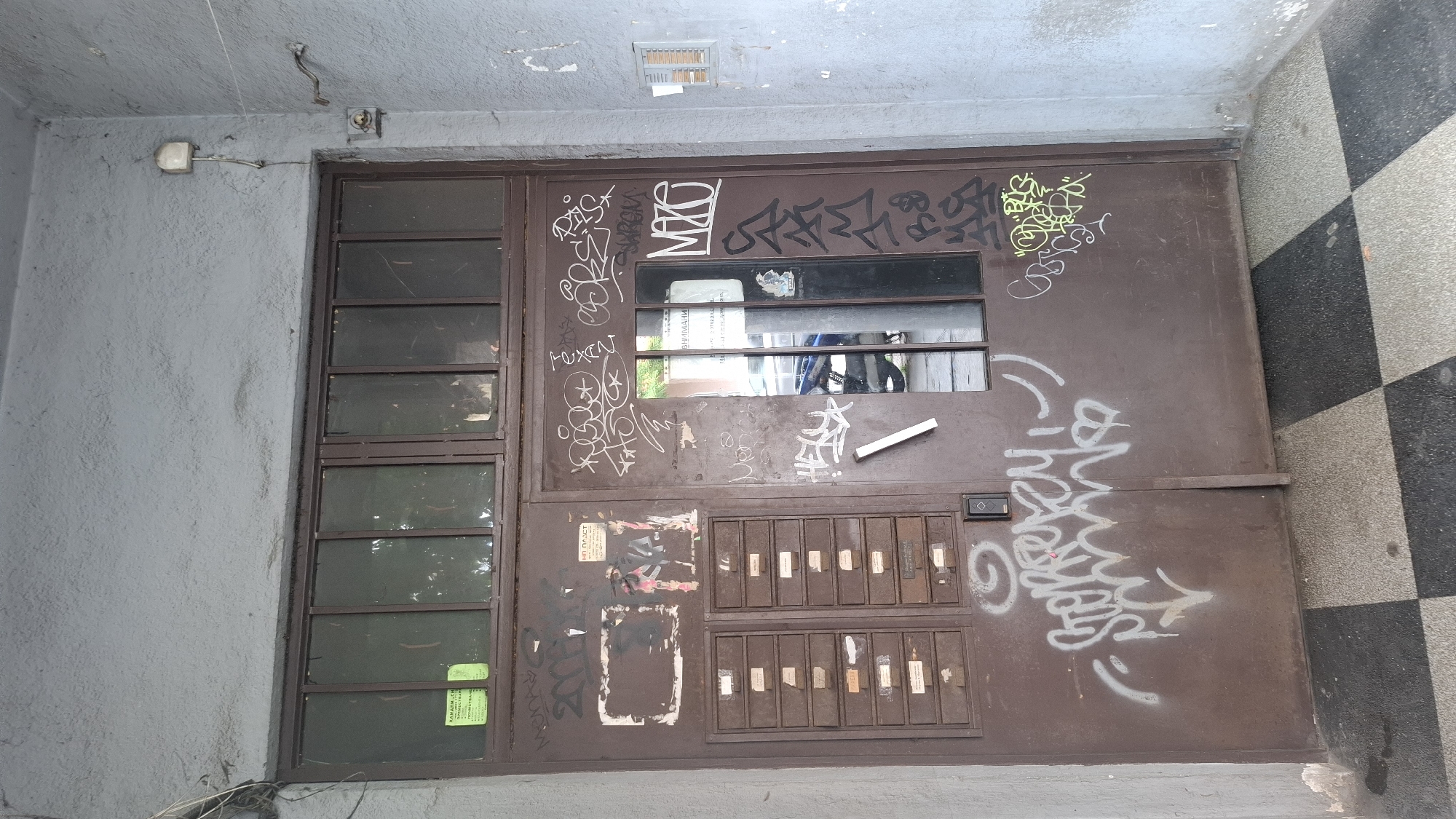 The image shows a close-up, eye-level view of a brown metal door with a glass panel at the top and a row of mailboxes on the left side. The door and the surrounding gray wall are covered in graffiti. The door itself is weathered and shows signs of wear. The glass panel at the top of the door has several panes and some stickers are visible on the interior. Below the mailboxes, there is a digital lock panel. On the right side of the door, there is a vertical glass panel that reflects the outside. The floor in front of the door has a black and white checkered pattern. The graffiti includes various tags and larger artistic pieces in white, black, and green spray paint. The overall environment appears to be an entryway or hallway in an urban setting, consistent with a location in Sofia, Bulgaria, given the context. There are no people or animals visible in the frame.