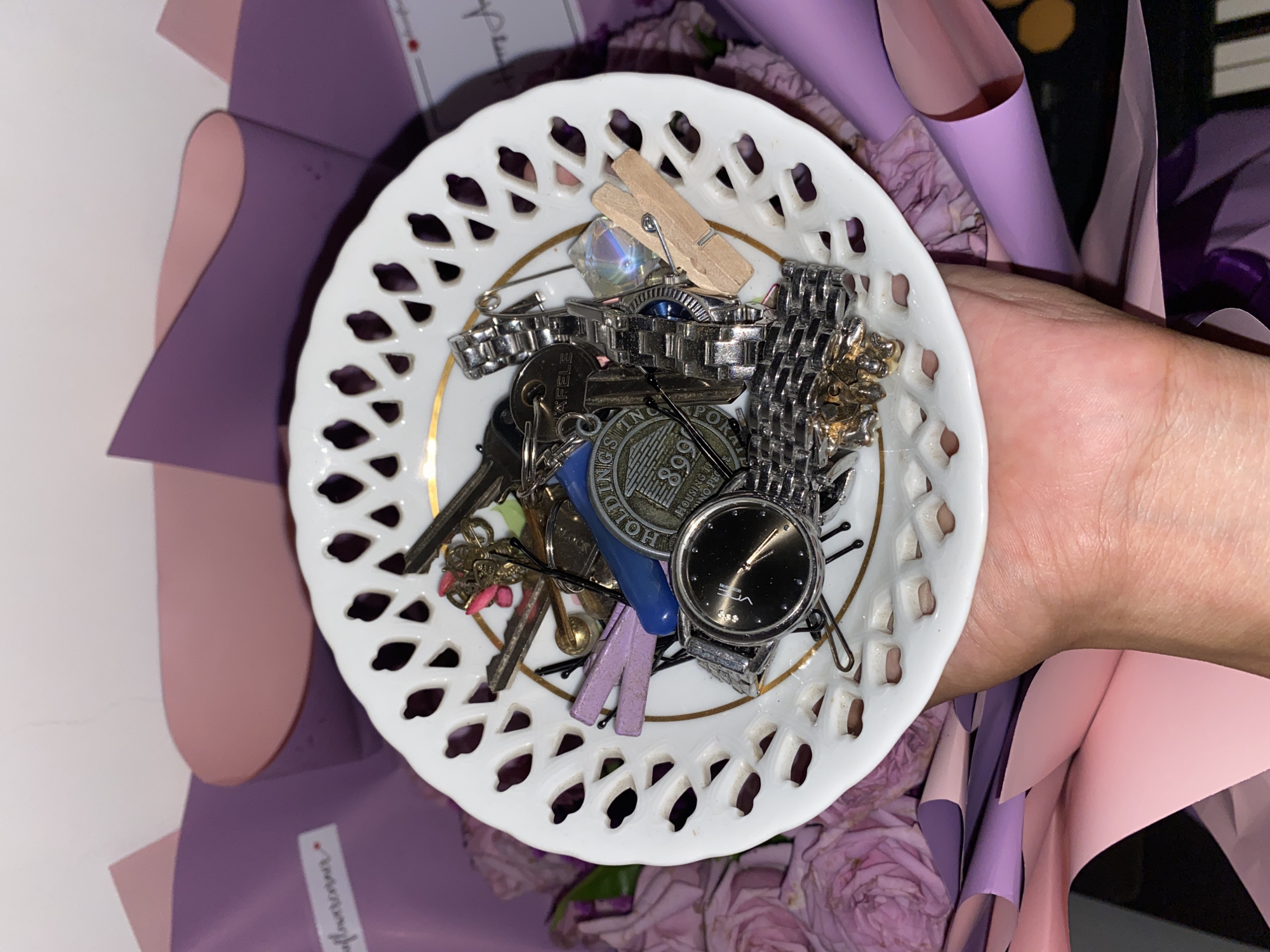 A hand holds a small, white, pierced porcelain dish with a gold rim. The dish contains an assortment of objects, including keys, a silver wristwatch, a coin, a safety pin, a small wooden clothespin, and other metallic trinkets. The dish is surrounded by a bouquet of flowers wrapped in purple and pink paper. The environment includes what appears to be a white surface on one side and a dark background on the other. There is no information to suggest the photograph was taken in Davao City, Philippines.