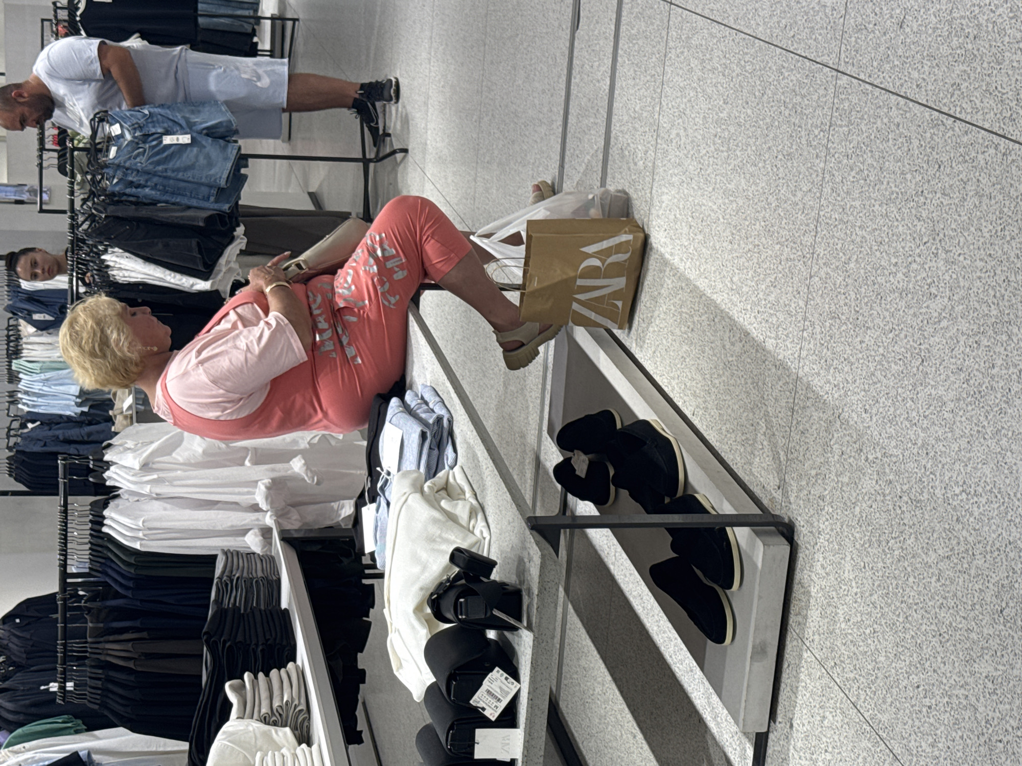 This image captures an interior scene within a Zara clothing store, located in Plovdiv, Bulgaria. The floor is covered with light gray speckled tiles featuring visible grout lines.
In the left foreground, a middle-aged woman with blonde hair is seated on a light gray display bench. She is dressed in a light pink t-shirt layered under a coral-pink sleeveless jumpsuit or capris with a silver textual design on the lower legs. Her gaze is directed downwards towards a light-colored handbag held in her lap. She wears light-colored, chunky-soled sandals. Adjacent to her feet are a brown paper Zara shopping bag and a clear plastic shopping bag.
In the mid-ground to the right, a man with a bald or closely shaven head and a beard stands, examining a pair of blue denim shorts. He is attired in a light blue t-shirt with a Nike logo and light blue shorts with a white Nike logo. His footwear consists of dark athletic shoes with white soles.
A third individual, a young woman with dark hair styled in an updo, is partially visible in the background, positioned behind a clothing rack between the other two individuals. She appears to be wearing a dark top and is looking towards the right side of the frame.
The store's inventory is displayed on multiple metal clothing racks: dark garments (black, navy, green) are on the left, a selection of black and blue denim shorts are centrally located, and white long-sleeved shirts are on the right. A display unit behind the seated woman features neatly folded stacks of clothing in white, light gray, and blue stripes, alongside black small bags or wallets with attached price tags, some indicating "ZARA" branding. A lower shelf beneath this unit showcases multiple pairs of black loafer-style shoes with light-colored soles. The scene is illuminated by overhead artificial lighting.