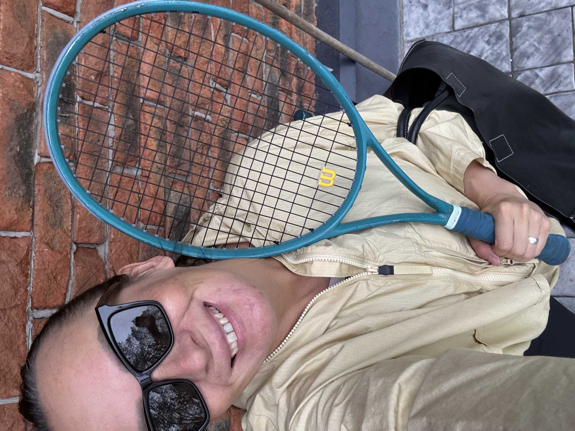 In a selfie taken outdoors, a smiling woman wearing sunglasses and a light-colored jacket holds a turquoise tennis racket. The background consists of a red brick wall and patterned paving stones. A black tote bag is visible on the right side of the frame. The image does not contain any information indicating the location in Chaikovsky, Russia.