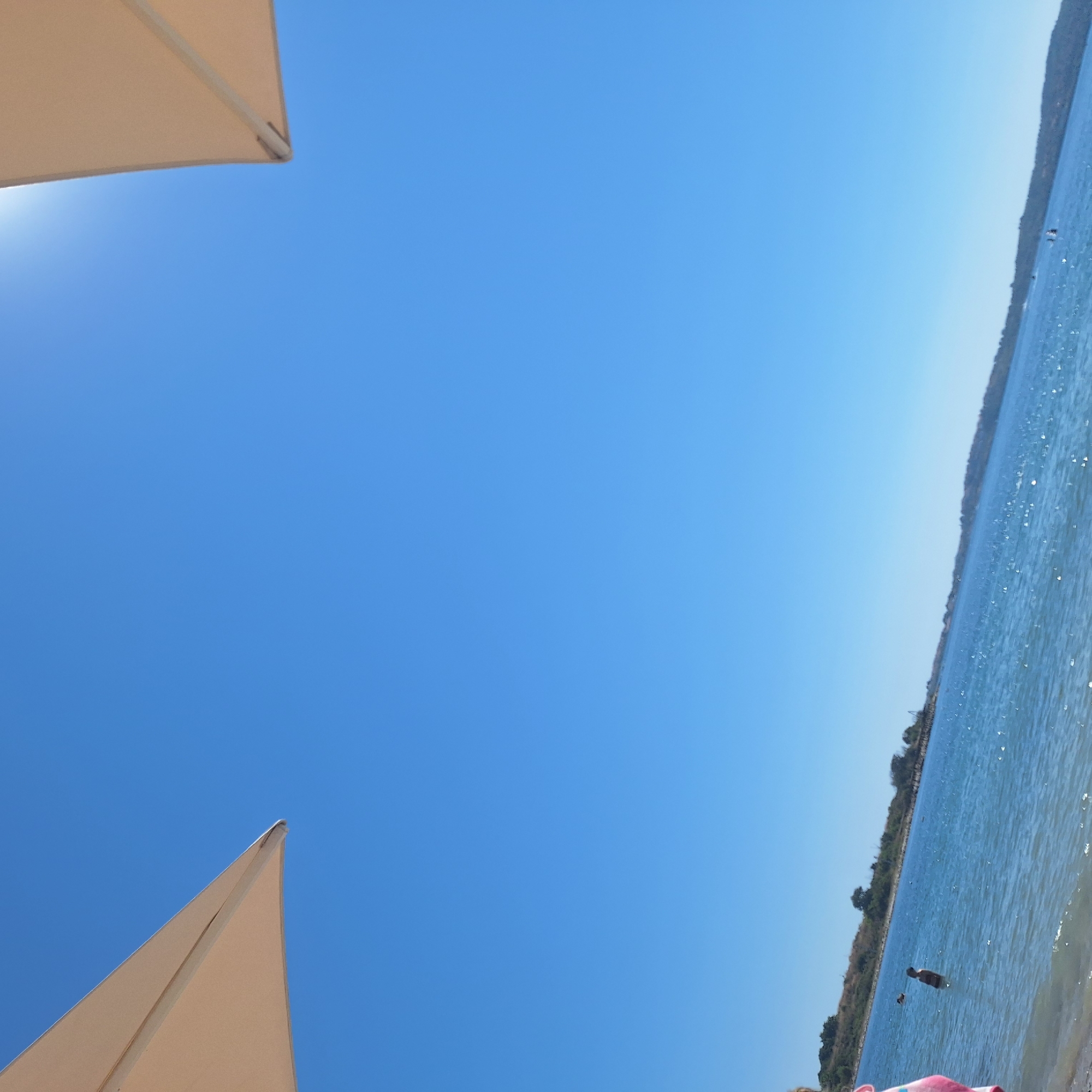 This is a medium shot, low-angle view of a clear blue sky and a body of water. In the foreground, on the left, a portion of a beige beach umbrella is visible, angled towards the upper left corner. On the right, another part of a similar umbrella is visible, angled towards the upper right corner. The water is a deep blue with some light reflection on its surface. In the middle ground, a person in dark swimwear is standing in the water. Further out, other swimmers are visible. The background shows a tree-lined shore. The location is the sea near Chernomorets, Bulgaria. The weather is sunny.