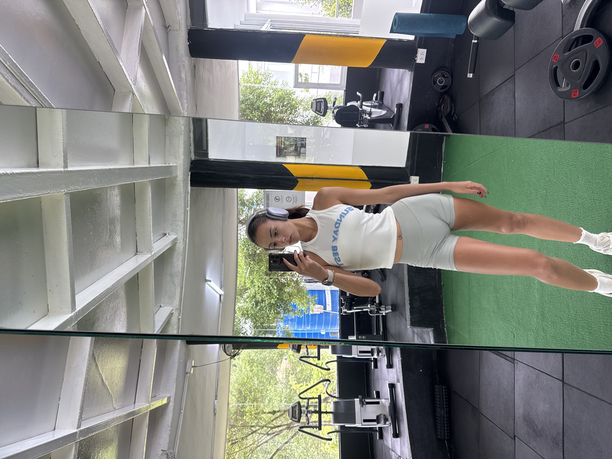 A female stands in a gymnasium, taking a mirror selfie with a smartphone. She is wearing a white tank top, light green shorts, white socks, and white sneakers. She has headphones around her neck. The gymnasium has black and yellow painted pillars, various exercise machines including a stationary bike, and black and green flooring. The location appears to be in Kathu, Thailand, as scenery and lighting suggest a tropical environment.