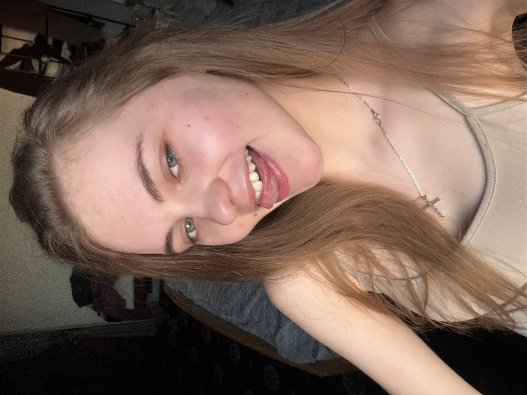 A young woman with long, straight, light brown hair and light blue eyes is shown in a selfie. She is wearing a light-colored tank top and a silver necklace with a cross pendant. Her mouth is open, and her tongue is sticking out slightly. She has a mole on her cheek and several small red blemishes on her face. She is indoors, with a blurred background that includes a chair and some dark fabric. The image is of a close-up of her face and upper body. There is no identifiable information to suggest the location is Dnipro, Ukraine.