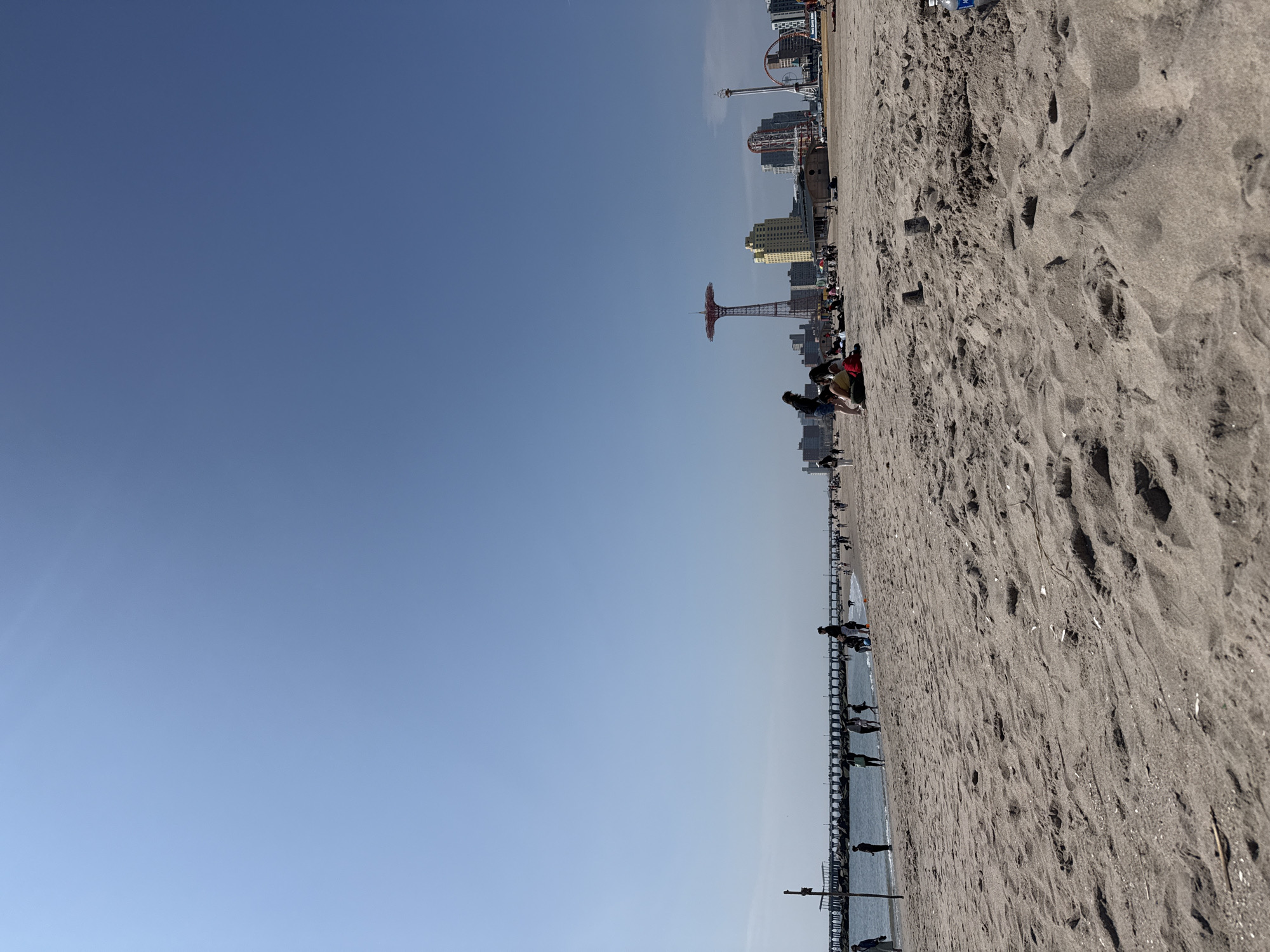 The image depicts Coney Island Beach in Brooklyn, New York City. The scene captures a clear blue sky covering half the image's space, juxtaposed against the beach area with brown sand. In the distance, the iconic parachute jump tower and other amusement park rides are visible. People populate the beach, mostly in small groups, engaging in recreational activities like sunbathing or strolling along the pier. The pier stretches into the ocean, appearing to be made of dark wood. The overall setting suggests a sunny day at the beach, typical of Coney Island.