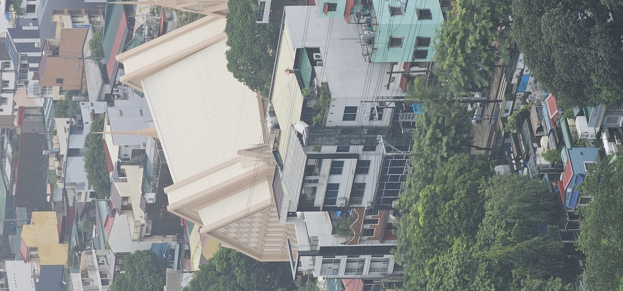 This is an aerial shot of the Araneta City in Quezon City, Philippines, a mixed-use development featuring residential, commercial, and entertainment spaces. The image shows a dense urban landscape with various buildings of different architectural styles and heights. Prominently featured is a large, modern building with a distinctive cream-colored, angular roof, likely a church or convention center, characterized by its sharp peaks and geometric patterns on its facade. Surrounding it are lower-rise buildings, some appearing residential with balconies and windows, while others seem to be commercial establishments with glass storefronts. Lush green trees are interspersed throughout the scene, providing a natural contrast to the concrete structures, suggesting the presence of parks or well-landscaped areas. The overall impression is one of a bustling city with a blend of modern architecture and pockets of greenery.