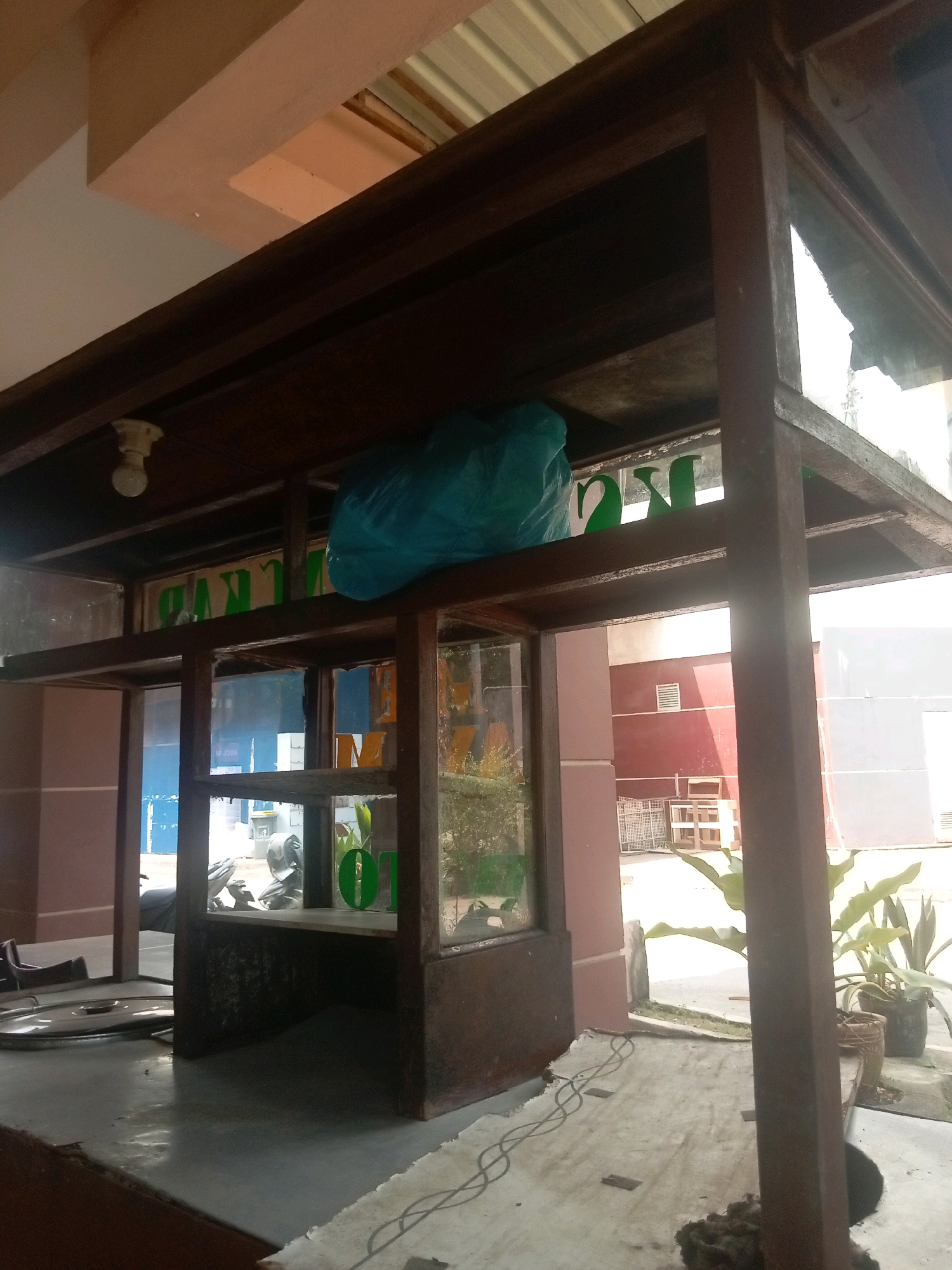 The image shows a street-side establishment with a dark brown wooden frame structure, possibly a food stall or kiosk, in Batam, Indonesia. The structure has multiple levels and glass panels. On the upper level, a turquoise plastic bag is placed on a wooden shelf. A lightbulb is visible on the ceiling of the upper section. Green lettering, partially obscured, is affixed to the wooden frame behind the glass panels, indicating some form of signage.
The lower section of the structure features glass-enclosed displays. Inside one of these displays, plants are visible. Outside, a motorcycle is parked near the structure, and plants are seen in pots near the right edge of the frame. The ground in front of the structure is a concrete surface covered with a worn, light-colored sheet or tarp, with some visible electrical wiring. In the background, a building with reddish-brown and pale pink sections is visible, along with some outdoor furniture and what appears to be fencing. The lighting suggests it is daytime, with sunlight casting shadows.