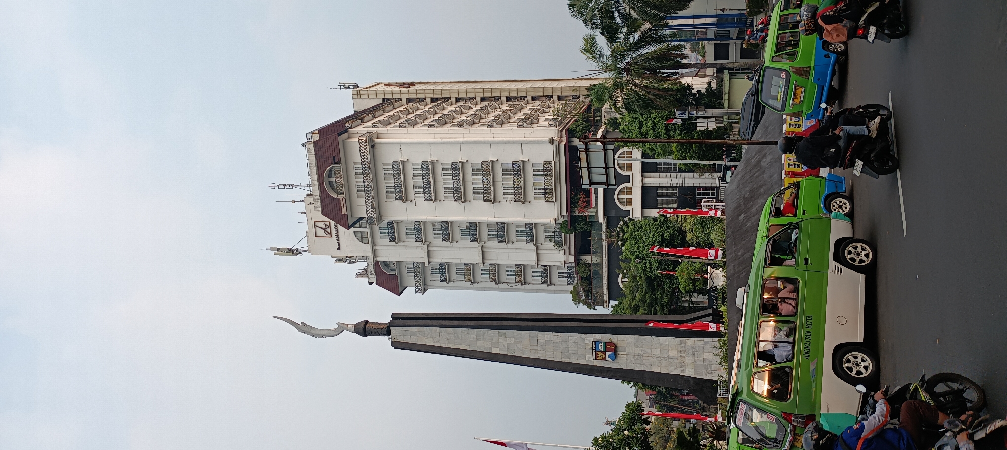 The image captures a street scene in Bogor, Indonesia, dominated by the "Tugu Kujang" monument on the left and a tall white building with a red roof, identified as the Royal Amaroossa Hotel Bogor. The Tugu Kujang, a significant landmark in Bogor, features a stylized kris (a traditional Indonesian dagger) atop a tapering, obelisk-like structure adorned with stone panels displaying emblems.
In the foreground, a busy intersection is visible with several vehicles. Two green and white minibuses, labeled "ANGKUTAN KOTA," are prominent. The one on the left is filled with passengers, while the one on the right is also in motion. Several motorcycles are also present, with riders navigating through the traffic.
The environment appears to be an urban setting with a clear sky. Red flags are visible, possibly part of a local celebration or event, planted along the roadside and near the monument. Palm trees and other greenery add to the tropical atmosphere of the scene. The overall impression is of a bustling city environment with distinct cultural elements.