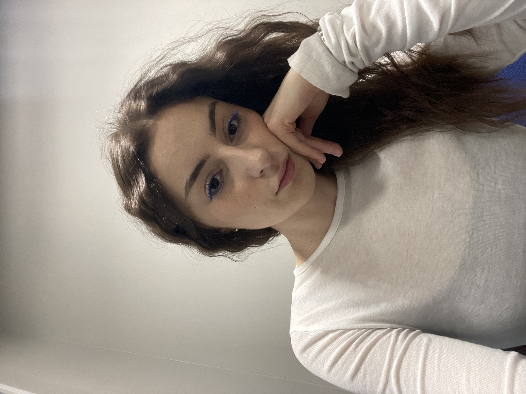 A young woman poses for a selfie. She has long, wavy brown hair and is wearing a long-sleeved white shirt. Her hand is resting on her face. She is wearing blue eyeliner. The background is a plain white wall. There is no information about the location or Moscow, Russia in the image.