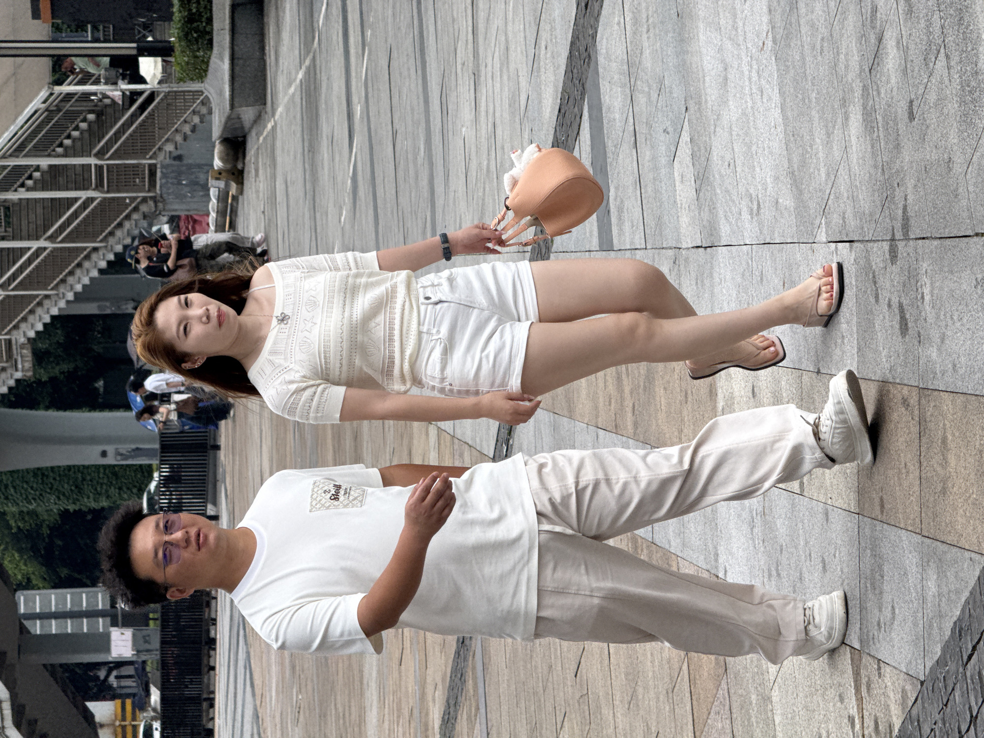 In Chongqing High-tech Zone, China, a man and a woman are standing on a tiled walkway. The woman, with red hair, wears a white short-sleeved top, white shorts, and tan sandals. She holds a tan purse in her right hand. The man, with short dark hair and glasses, wears a white t-shirt and light tan pants with white sneakers. They are in front of a metal staircase, with other people visible in the background.