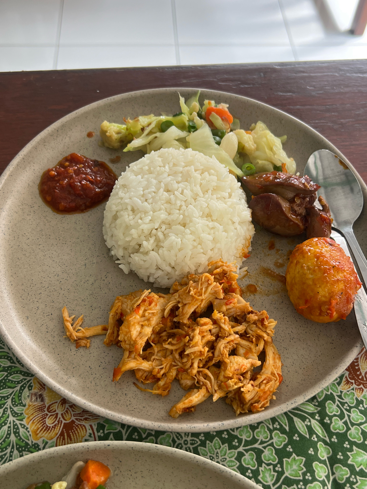 A close-up overhead shot shows a meal served on a speckled grey plate, resting on a green cloth with a floral pattern. The meal includes a mound of white rice, shredded chicken in a reddish sauce, a fried egg coated in sauce, a dollop of red chili paste, and a medley of cooked vegetables including cabbage, carrots, and what appears to be cauliflower. Several pieces of dark brown, glossy meat, possibly chicken liver or gizzards, are also on the plate. A silver spoon is partially visible on the right side of the plate. The setting appears to be a table, with a wooden surface visible in the upper portion of the image and a tiled wall in the background. This meal is likely from a restaurant in Kuta, Indonesia, a popular tourist destination known for its diverse culinary offerings.