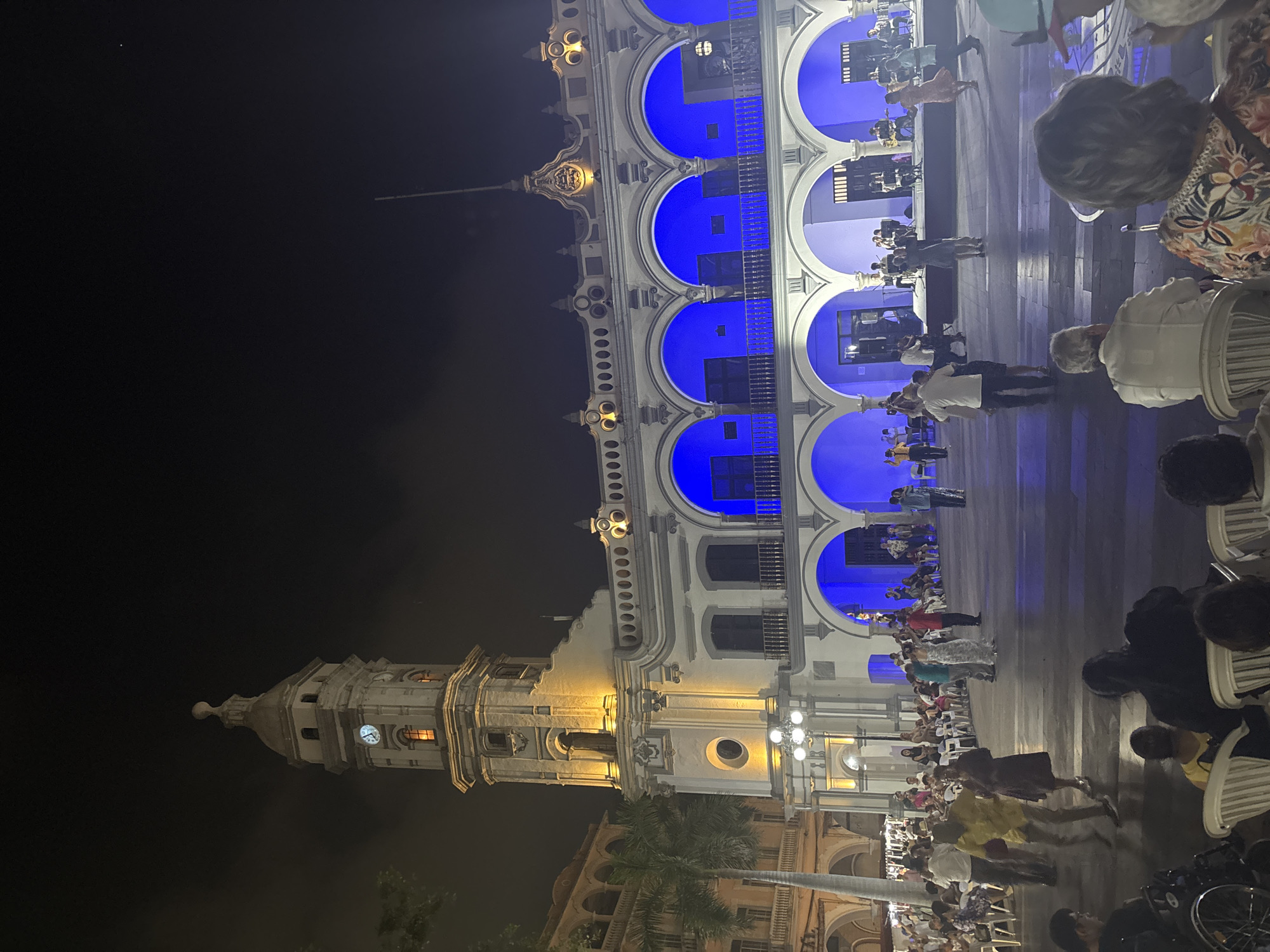Captured at night, the image shows a grand building in Veracruz City, Mexico. The building is illuminated with a combination of yellow and blue lights, highlighting its architecture and architectural details like the arches on the upper level. Numerous people are gathered in what appears to be a public space in front of the building. Some are seated, watching or listening to something, while others are standing or walking around. There appear to be musicians performing. The area is well-lit, allowing details of the scene to be clearly visible despite the darkness.