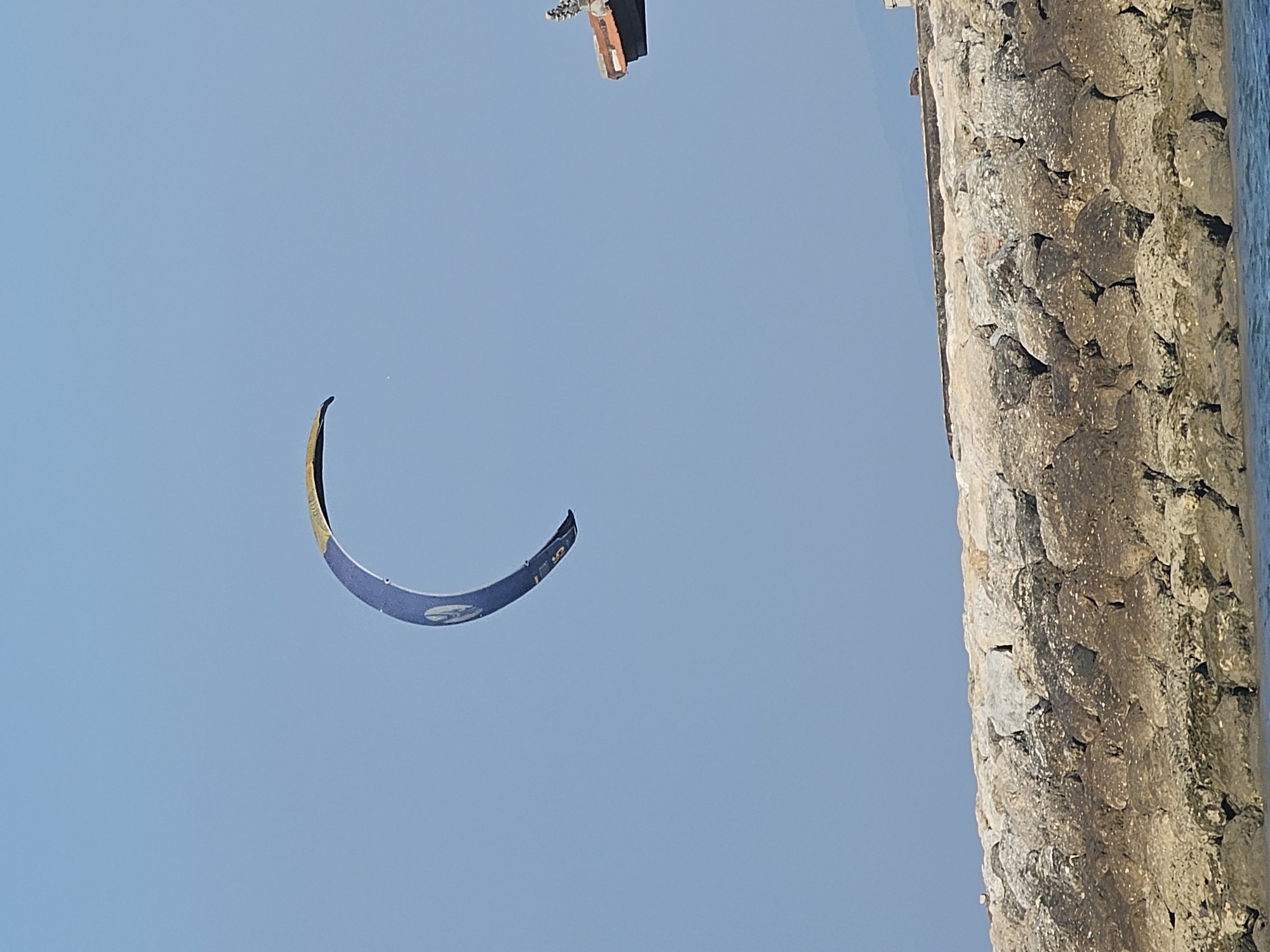 A blue and yellow kite is in the sky above a rocky breakwater. The kite is curved in a crescent shape, with its tip pointing towards the right. The breakwater is made of large, irregular stones, and a portion of a building with a red roof is visible on the right side of the image, behind the breakwater. The sky is clear and blue, with a faint hint of mountains in the distance. The water is calm and blue. The scene is likely a beach or coastal area in Denpasar, Indonesia, a popular spot for water sports and recreation.