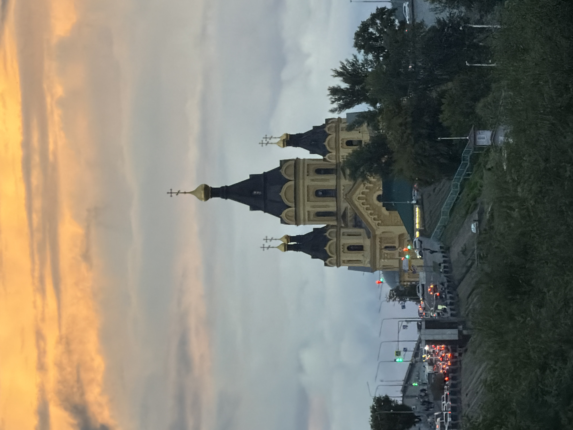 The image depicts a view of the Alexander Nevsky Cathedral in Nizhny Novgorod, Russia, as seen from a distance. The cathedral, with its distinctive yellow walls and dark blue onion domes topped with golden crosses, dominates the upper center of the frame. The sky above is filled with dramatic, cloudy formations, featuring streaks of warm orange and soft grey. In the foreground and middle ground, a dense arrangement of green trees and foliage obscures the lower portion of the scene. Below the cathedral, a busy street with a traffic jam is visible, indicated by numerous cars with illuminated brake lights and headlights, and several traffic signals displaying red and green lights. The presence of streetlights and the illuminated cityscape suggest it is either dusk or dawn. A grey, stepped embankment with a green railing leads up towards the cathedral's foundation. The overall scene captures a twilight view of a prominent religious landmark and the bustling urban environment surrounding it.