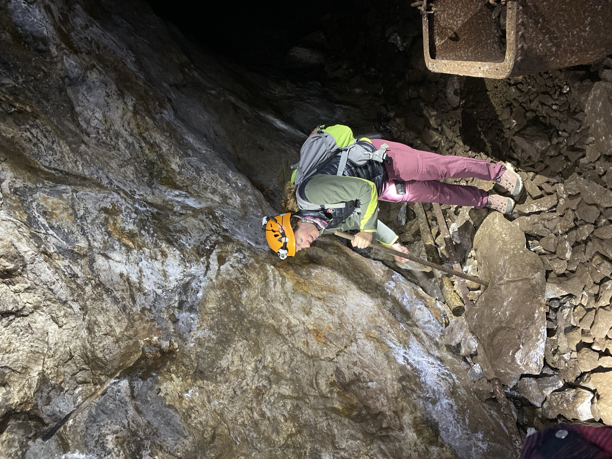An individual wearing caving or mining gear is navigating a dark, rocky underground passage, likely a cave or mine shaft in Les Chavannes-en-Maurienne, France. The person, appearing to be female, is equipped with an orange helmet featuring a prominent headlamp, a grey backpack, a dark padded vest over a light green long-sleeved top, and purple or maroon pants. They are wearing sturdy hiking or caving shoes.
The individual is positioned on what appears to be a rudimentary ladder or support structure, made of wooden or metal beams, and is holding onto one of its rungs with both hands. Their body is angled, suggesting they are either ascending or carefully descending a steep section of the passage. The headlamp casts a bright beam onto the immediate surroundings, illuminating the rugged and uneven rock surfaces of the cave walls and ground.
The environment is characterized by dark, damp-looking rock formations with varying textures and shades of grey, brown, and white, possibly mineral deposits. The ground below is covered in loose, jagged rocks and debris. In the upper right of the frame, a section of an old, rusted metal channel or pipe is visible, firmly attached to the rock ceiling, hinting at past mining operations. The overall scene conveys a sense of exploration or work in a challenging and confined subterranean space, illuminated solely by artificial light. No distinct text is visible on clothing or equipment.