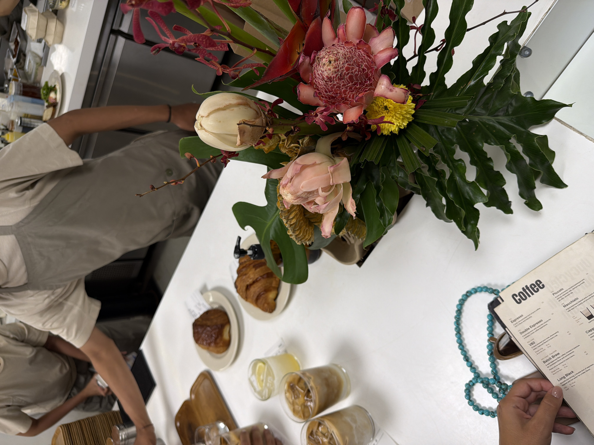 This high-angle photograph captures a scene at a café, possibly in Kuta Selatan, Indonesia. A floral arrangement featuring tropical flowers sits at the center of a white table. Around the floral arrangement are plates with pastries, including a croissant and a cake-like item, along with glasses filled with what appears to be iced coffee or tea and a lemon drink. Two individuals are partially visible: one serving drinks, and the other looking at a menu titled "Coffee", which lists various coffee drinks such as espresso, macchiato, and batch brew. A turquoise beaded necklace and sunglasses also lie on the table near the menu.