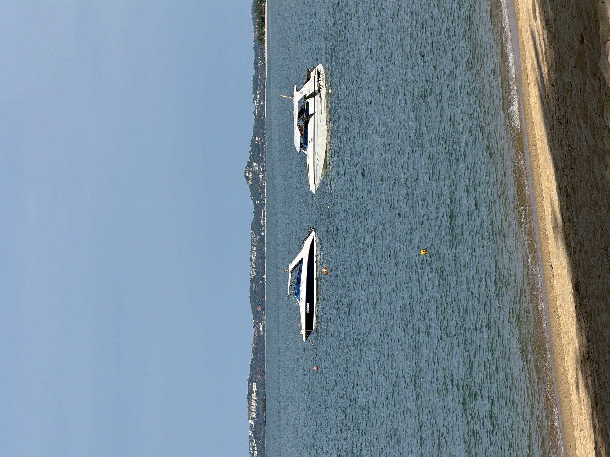 Two white speedboats are anchored in the turquoise water of the Gulf of Thailand near Baan Tai, Thailand. The boats are positioned side-by-side, with the boat on the left having a black hull. A distant, verdant coastline with buildings is visible on the horizon, under a clear, pale blue sky. In the foreground, a sandy beach is partially shaded by what appears to be palm fronds. Several buoys, one red and white, and one orange, are scattered in the water.