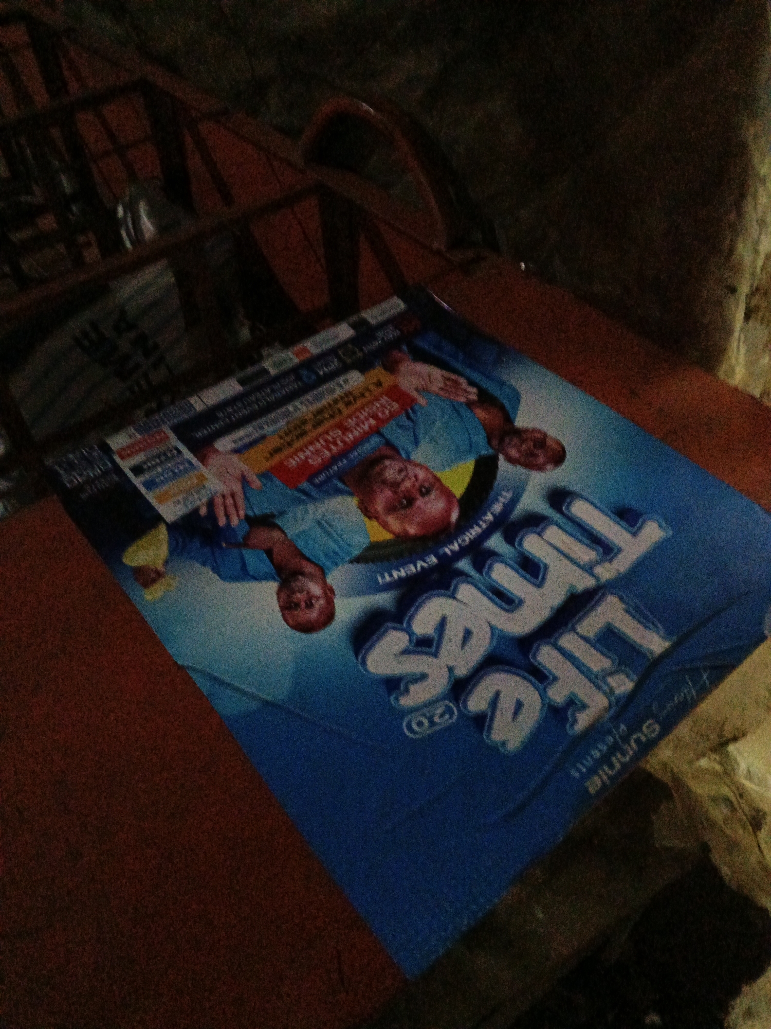 The image displays a poster for a theatrical event titled "LIFE TIMES" which features images of two men in blue shirts. One man, with a prominent smile and wearing a blue shirt, is positioned on the right side of the poster. Another man, also in a blue shirt, is shown on the left side, looking towards the viewer. The poster's design is predominantly blue with white lettering for the title. There are also various smaller text elements on the poster, some of which appear to be event details and sponsor logos. The poster is placed on a wooden surface, possibly a table, with a dark, textured background that suggests an indoor environment. The lighting in the image is dim, casting shadows and contributing to a grainy appearance of the photograph. The overall scene is a close-up of the poster, angled to capture its entirety, with the surrounding environment partially visible and out of focus. The location in the city of Bukuru, Nigeria, is not discernible from the visual information provided in the image itself.