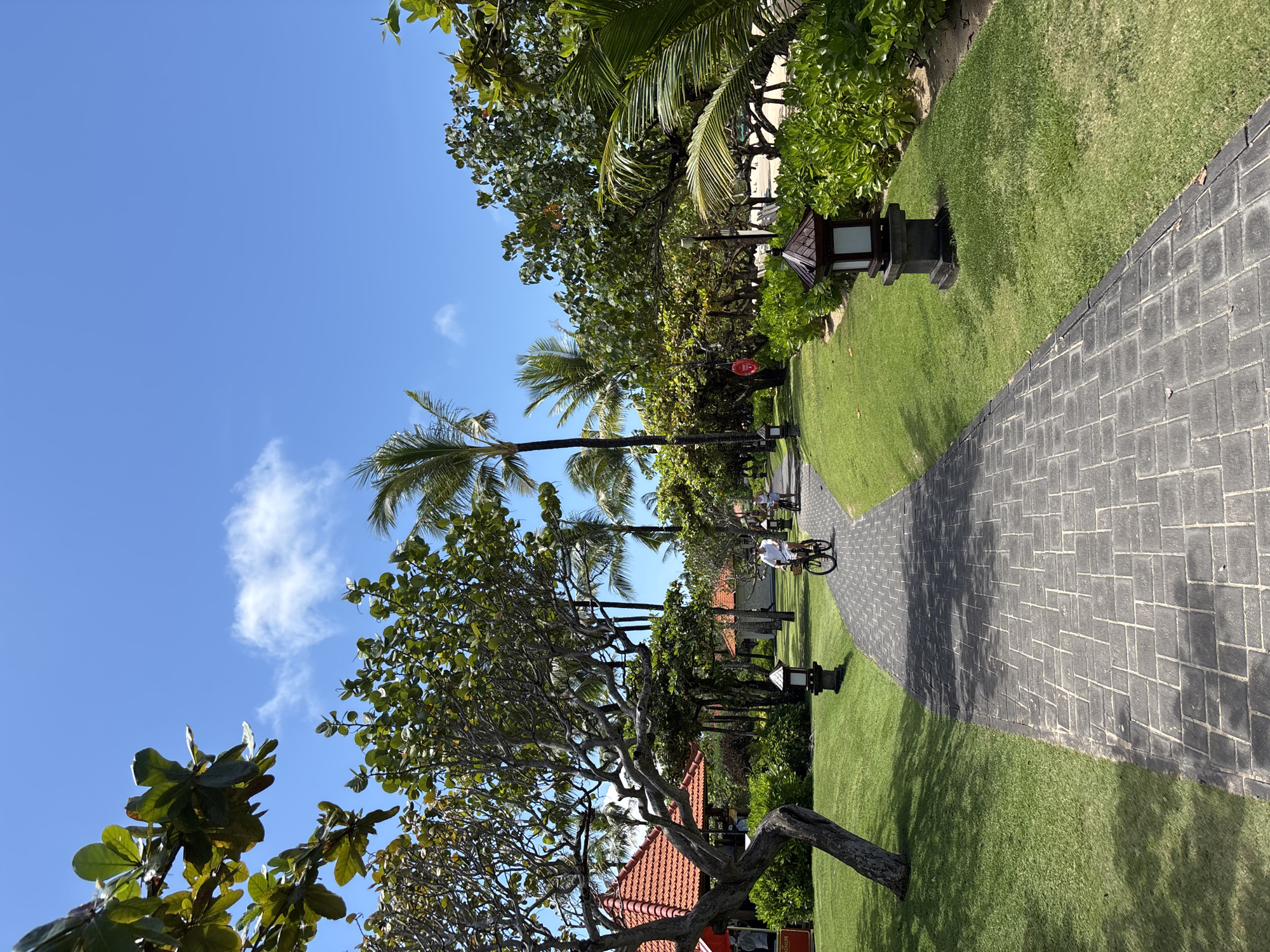 The image depicts a pathway in Nusa Dua, Indonesia. The pathway is made of dark grey interlocking brick tiles and curves through a grassy area lined with trees, including several palm trees. There are decorative lanterns along the path. A person is riding a bicycle on the path. Part of a building with a red-tiled roof is visible in the foreground. The sky is clear blue with a few scattered clouds.