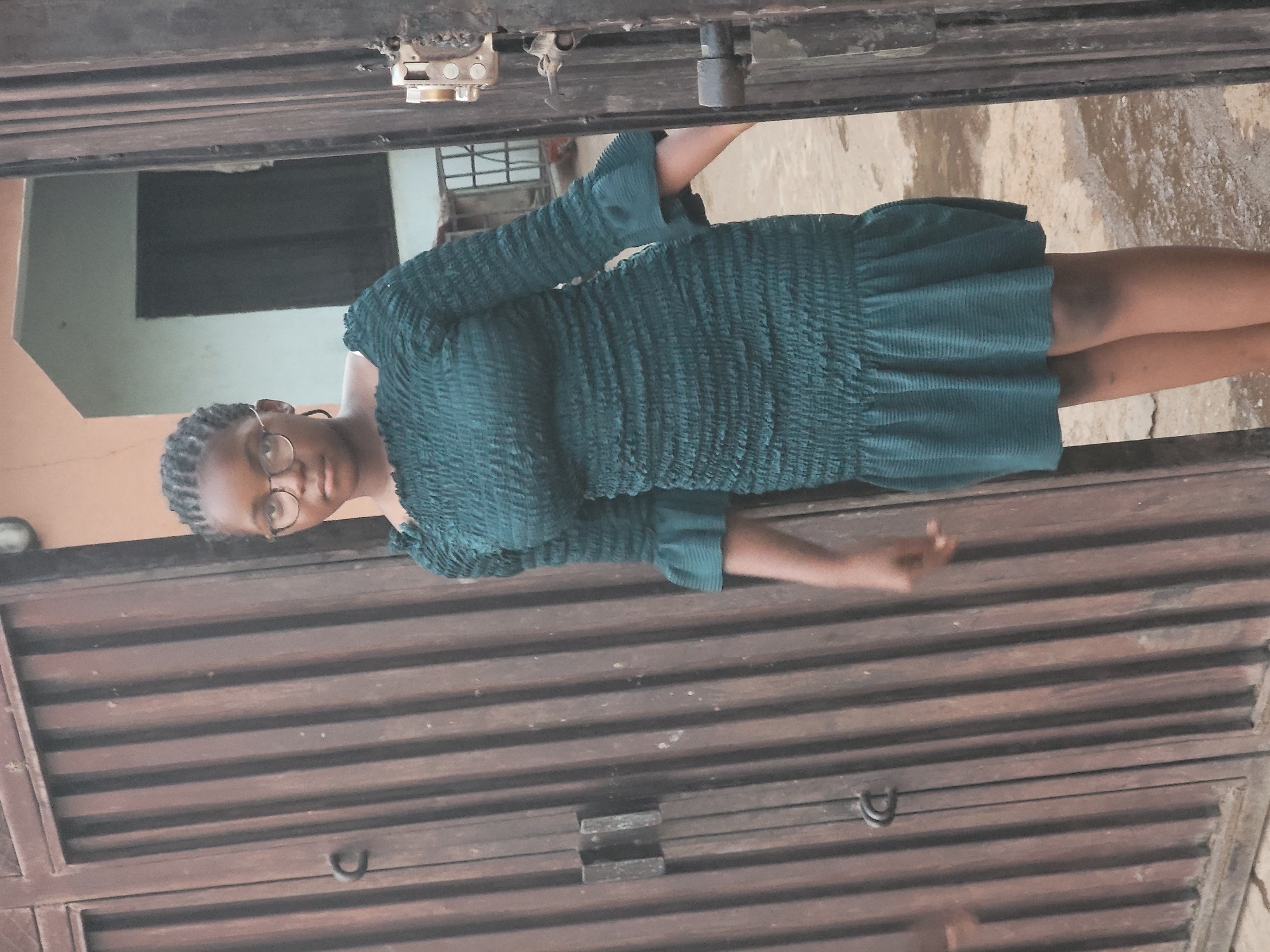 A woman wearing a green, textured dress and glasses is standing by a brown, horizontal-slatted gate. She appears to be either opening or closing the gate. The location appears to be outside, based on the background including a building exterior and pavement. The location is Sabon Tasha, Nigeria.