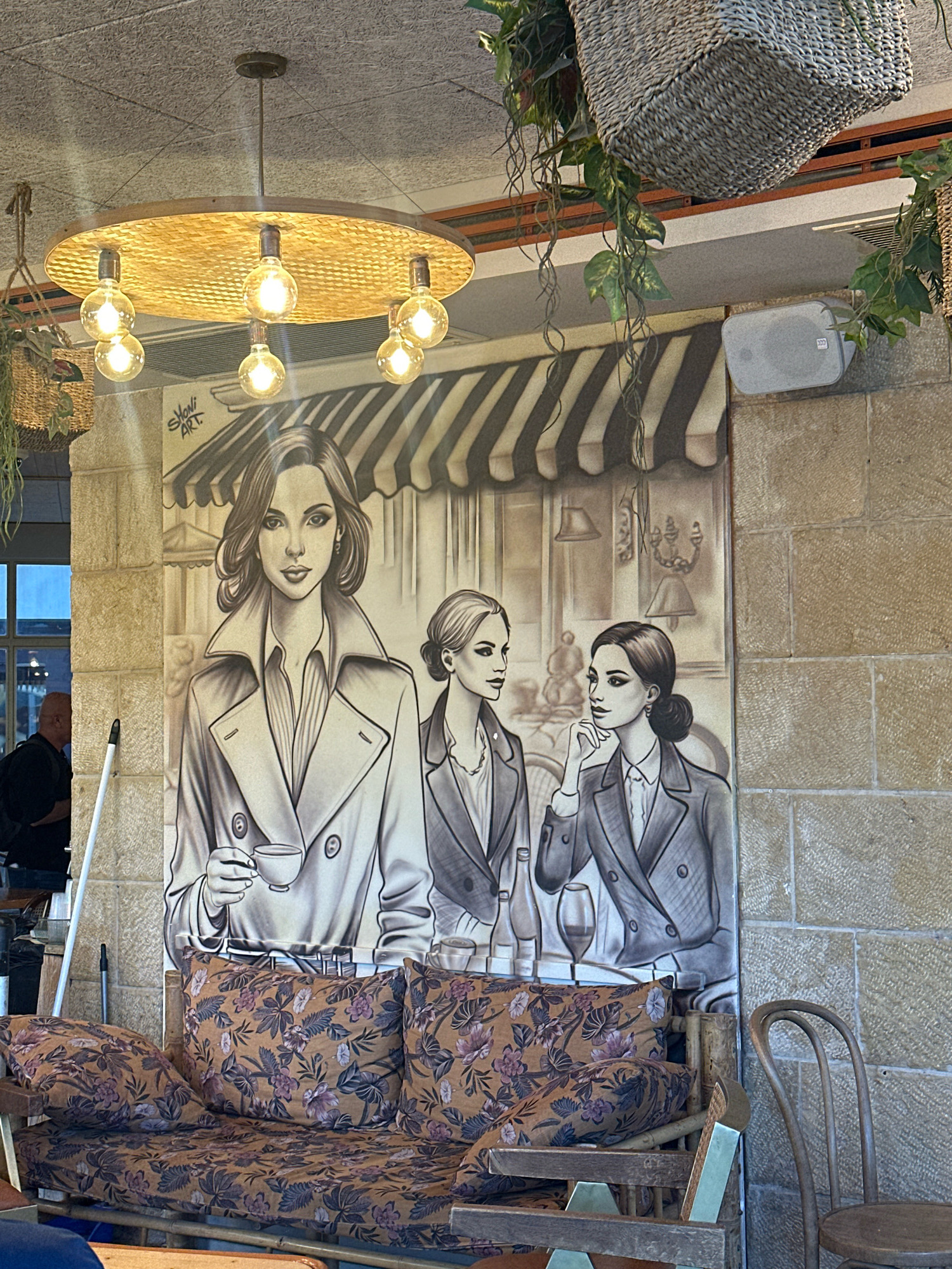 The image depicts an interior scene, likely a cafe or restaurant, in Jerusalem, Israel. The main focus is a large mural painted on a stone wall, featuring three women in a sketched, stylized manner. The mural also includes elements of what appears to be a Parisian street scene with awnings and cafe tables. In the foreground, a brown couch with floral patterned cushions is visible, alongside a wooden chair. Overhead, a light fixture with several exposed bulbs hangs from the ceiling, and decorative woven baskets with hanging plants are also present. The overall ambiance suggests a casual and artistic dining environment.