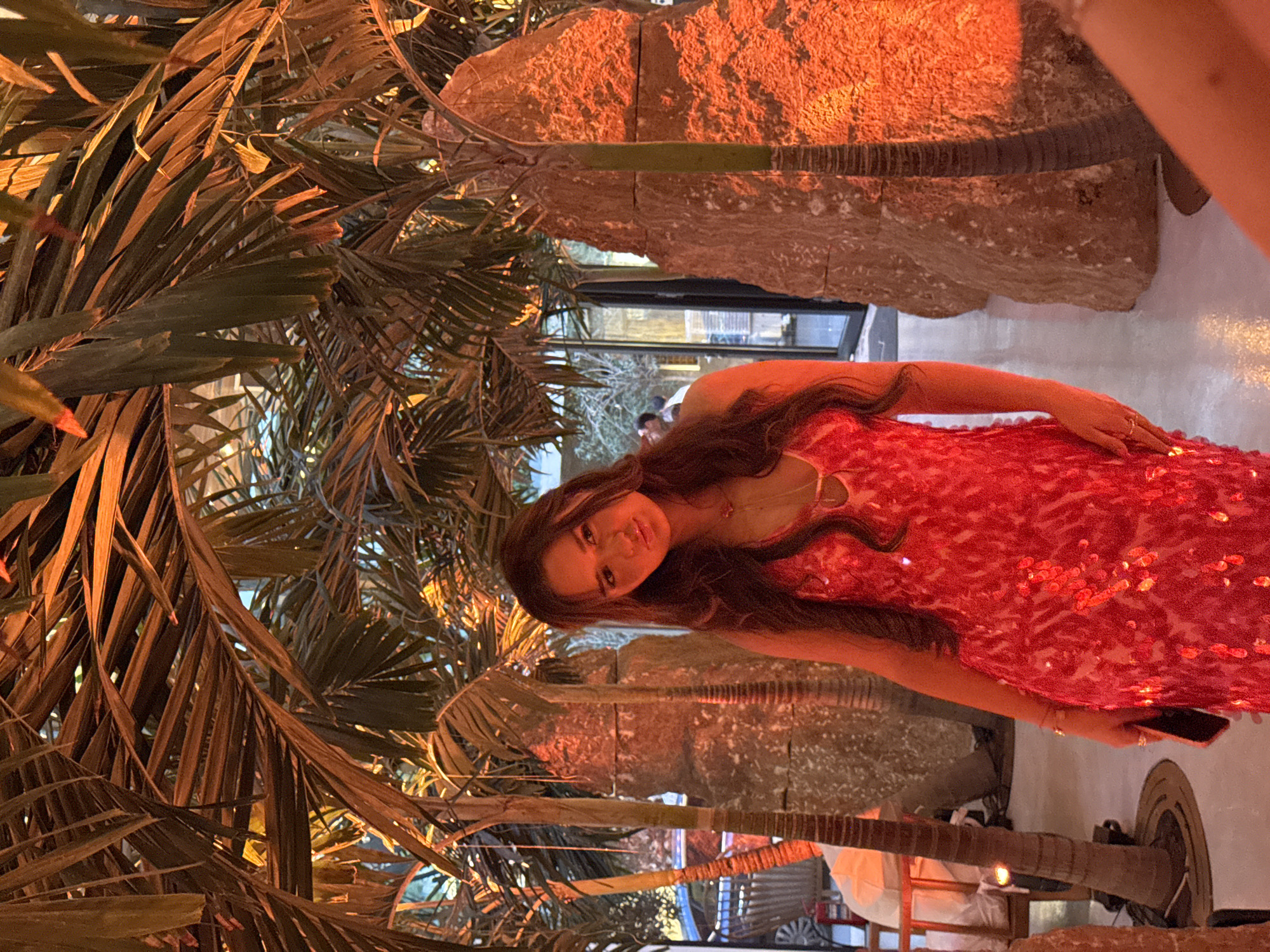 A young woman with long, dark wavy hair is standing in an indoor environment with a tropical theme, possibly a restaurant or event space, in Yerevan, Armenia. She is wearing a bright red, sequined dress with a low neckline and a necklace. Her expression is a pout. She holds a dark-colored smartphone in her right hand. She is positioned between several tall, rough-textured, light-colored rock formations and palm trees. The lighting is warm and orange, creating dramatic shadows. In the background, through a glass door, a few indistinct figures can be seen, suggesting the presence of other people and an outdoor area or another part of the building. A white piece of fabric is visible on the left side of the image, possibly part of another person or furniture. The floor appears to be smooth and reflective.