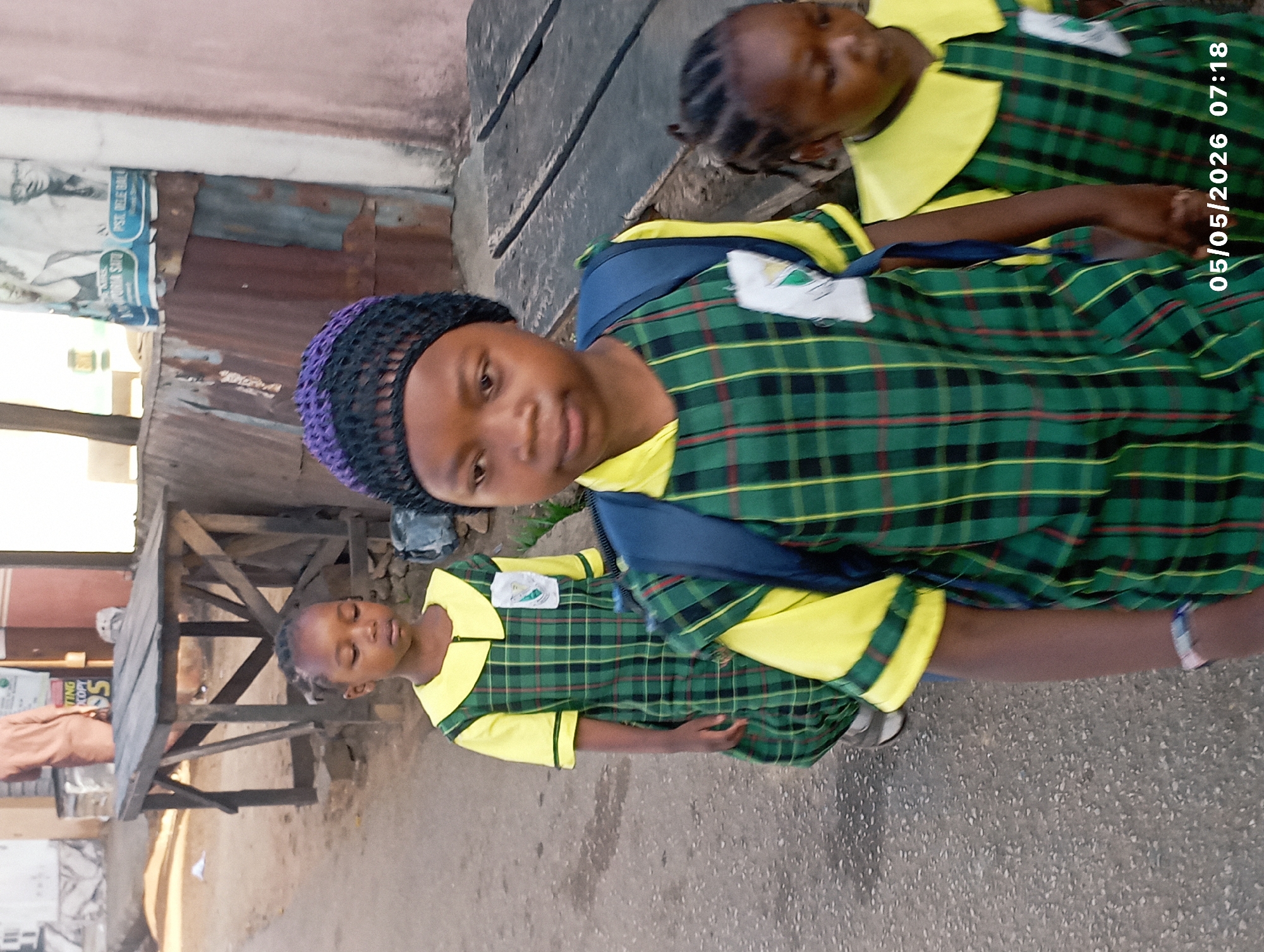 The image shows three young girls in green and yellow plaid school uniforms standing on a paved surface. One girl has a purple and black crocheted cap. All three are wearing backpacks. The background includes a weathered wooden table, a corrugated metal structure, and signage. The date "05/05/2026 07:18" is imprinted on the image. Given the architecture and environment, the location is likely an urban area, possibly within Jos, Nigeria.