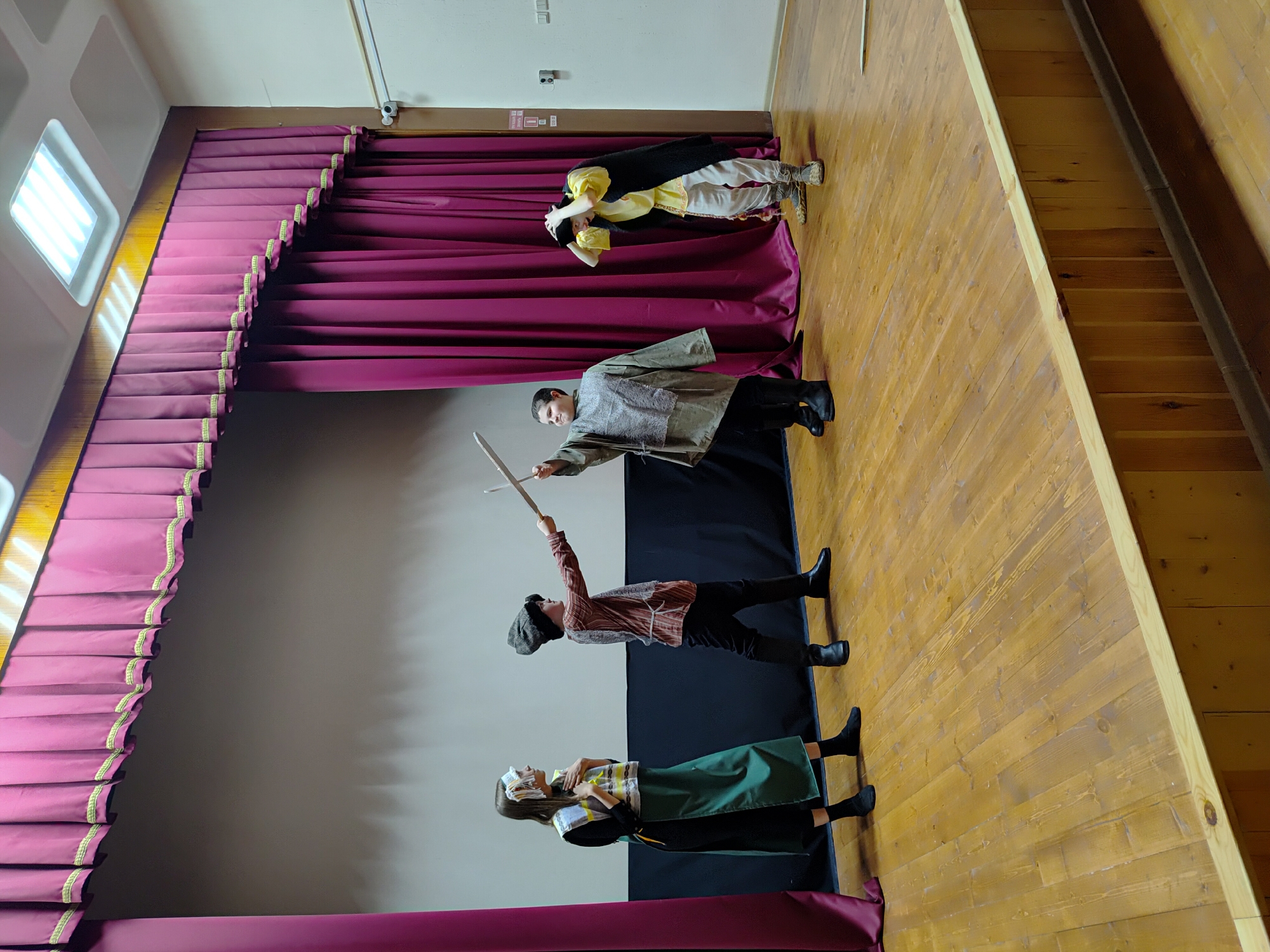 A stage performance is occurring. The stage has red curtains at the top and bottom and a plain wall behind the performers. Four individuals are on stage wearing costumes. The background and floor are unremarkable. No information suggests the location is in the city of Марковский, Russia.