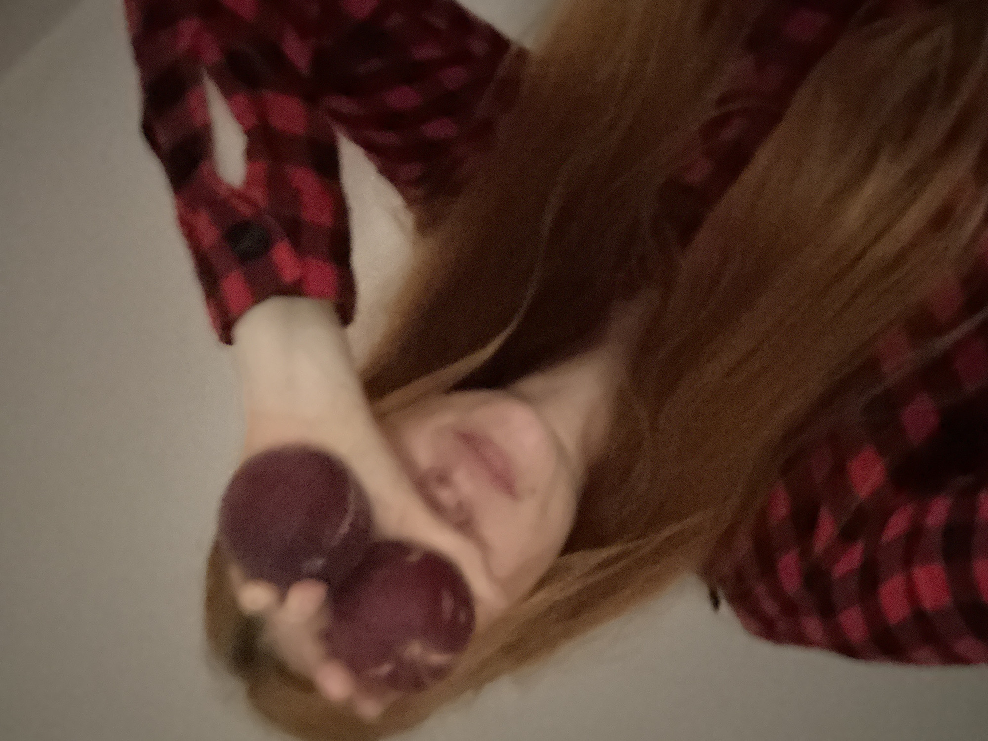 This media file presents an upward-angled, close-up shot of a person, creating the impression of a low-angle perspective looking up at them. The subject, likely female, has long, flowing light brown or auburn hair that extends across much of the frame. They are wearing a red and black plaid shirt, clearly visible on their arm and upper body.
The person's face is partially obscured by their hand, which holds two dark, reddish-purple, round objects in front of their eyes. These objects resemble small fruits, possibly plums. A subtle smile or smirk is visible on the person's lips, suggesting a playful or lighthearted mood. Their nose and forehead are visible above the held objects, with their eyes partially visible over them.
The background is a plain, light-colored surface, lacking any specific features or visual cues beyond its neutral tone. The lighting suggests an indoor setting. No specific time of day or weather conditions can be determined from the image. There is no visible text. The overall scene conveys a casual and candid moment, set in what is known to be Budva, Montenegro, though no specific geographical markers of the location are present in the visual content itself.