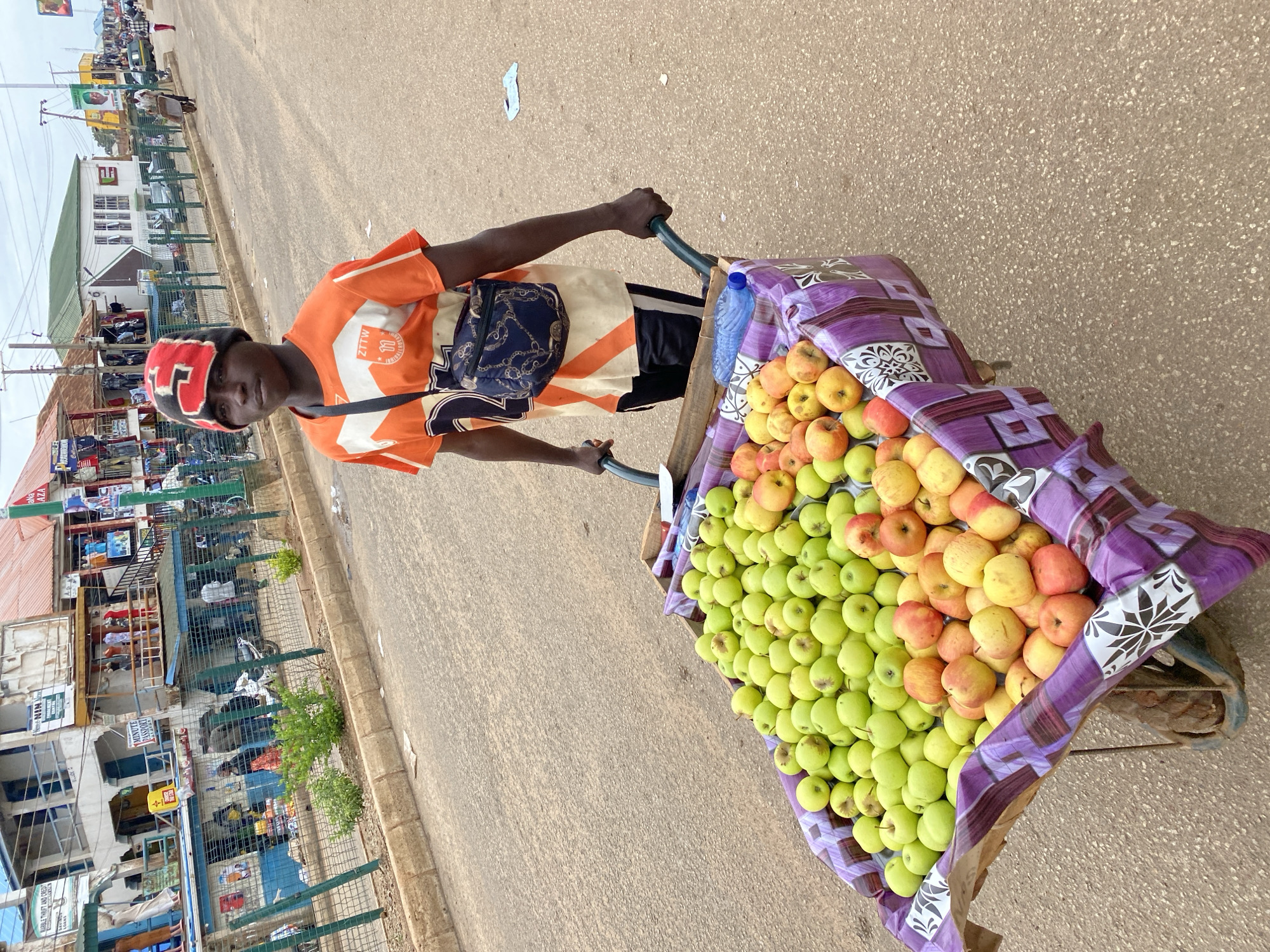 A young man, wearing an orange t-shirt and a patterned beanie, pushes a wheelbarrow full of apples down a paved street in Bukuru, Nigeria. The wheelbarrow is lined with a purple and white patterned cloth, and the apples are sorted into green and red varieties. Shops and buildings line the street, with some displaying clothing and other goods. Several other people are visible in the background, some walking and others standing near the shops. A green metal fence runs along the sidewalk. The sky is overcast, and the overall lighting suggests a daytime scene.
