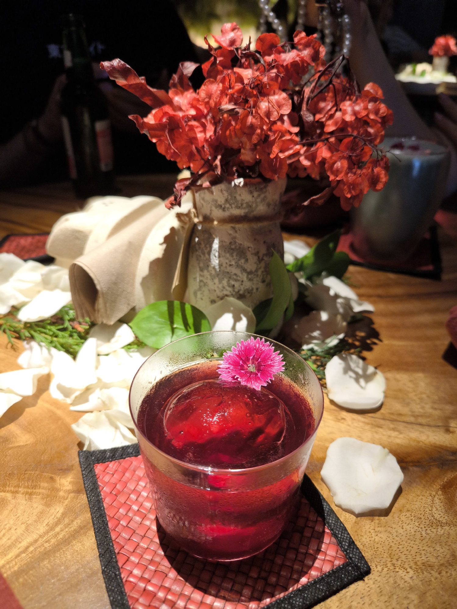 The image displays a close-up of a table setting, likely in a restaurant or bar in North Kuta, Indonesia. In the foreground, a clear glass holds a vibrant, dark red beverage with a large spherical ice cube and a small, bright pink flower used as a garnish. The glass rests on a textured red and black woven coaster.
The wooden table is adorned with numerous scattered white flower petals and sparse green foliage. In the midground, a decorative, rustic vase contains a prominent bouquet of dried, bright red flowers or foliage, which are the focal point of the upper half of the image. Behind these, a stack of rolled, light-colored napkins is visible.
The background is softly blurred. On the left, a person's hands and part of a dark green bottle, possibly beer, are indistinctly visible. On the right, another person's arm and a light-colored cup are partially obscured. The lighting creates distinct shadows from the flower petals on the table, suggesting an indoor setting with focused illumination, possibly during the evening. The overall scene conveys a decorative, possibly romantic or celebratory, atmosphere. No visible text is clearly discernible.