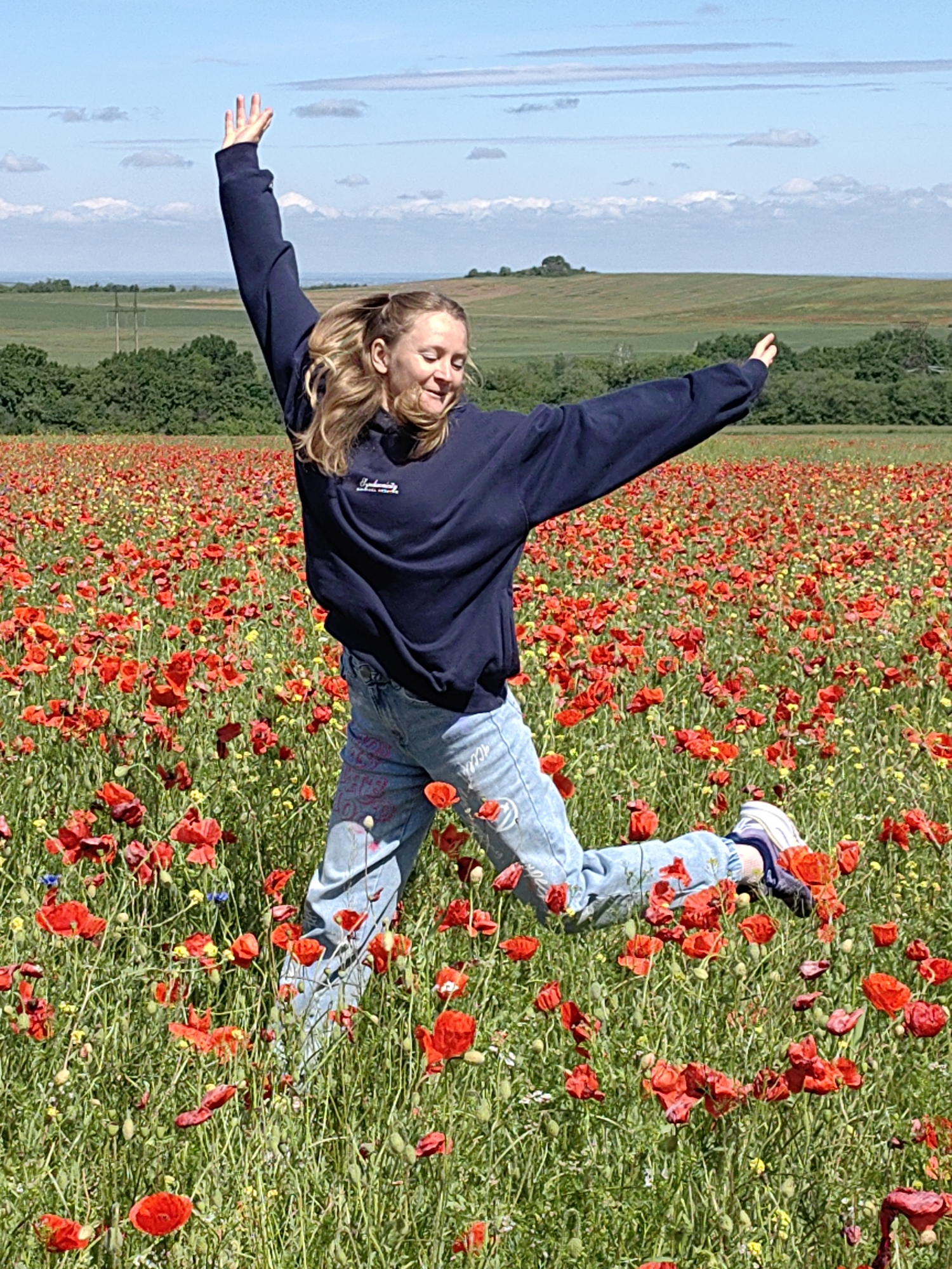 In a field of red poppies in Ароматновское сельское поселение, Ukraine, a young woman with blonde hair in a ponytail is jumping in the air with her arms outstretched. She is wearing a navy blue sweatshirt and blue jeans with writing on the legs. Her right foot is forward, wearing a blue and white sneaker. The background consists of green trees and rolling hills under a bright blue sky with some clouds.