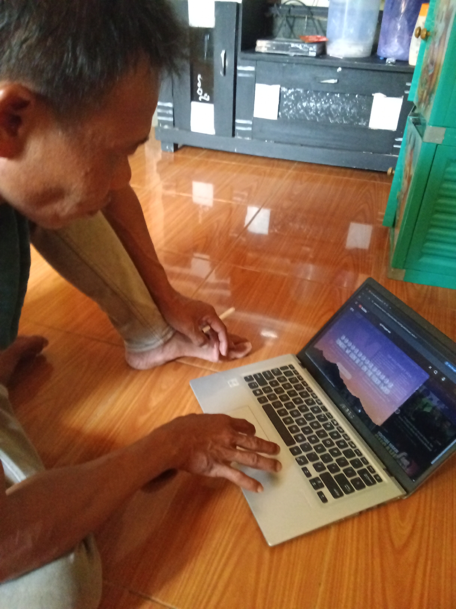 A person is seen from the side, sitting on a wooden floor and looking at a laptop. The person has dark hair and is wearing a dark shirt and light-colored pants. Their left leg is bent, and their right leg is extended. Their left hand is resting on the laptop's keyboard. The laptop screen displays a webpage with text and images. In the background, a dark cabinet with white accents is visible, along with some containers and a green cabinet. The floor is made of polished wood planks. This scene appears to be indoors, in a residential setting, possibly in the city of Subang, Indonesia.